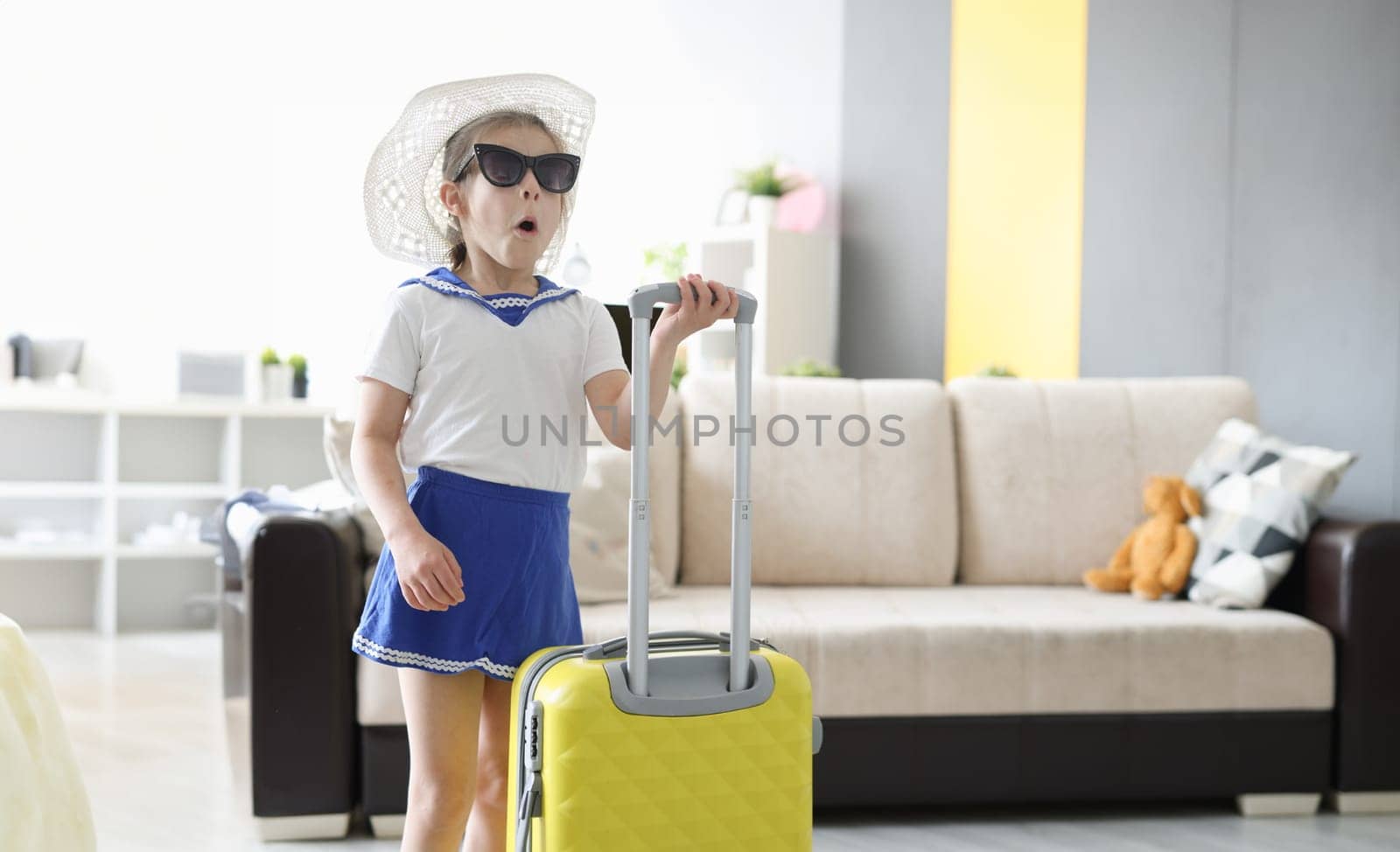 Surprised little girl in sunglasses and hat standing with suitcase in her hands at home by kuprevich