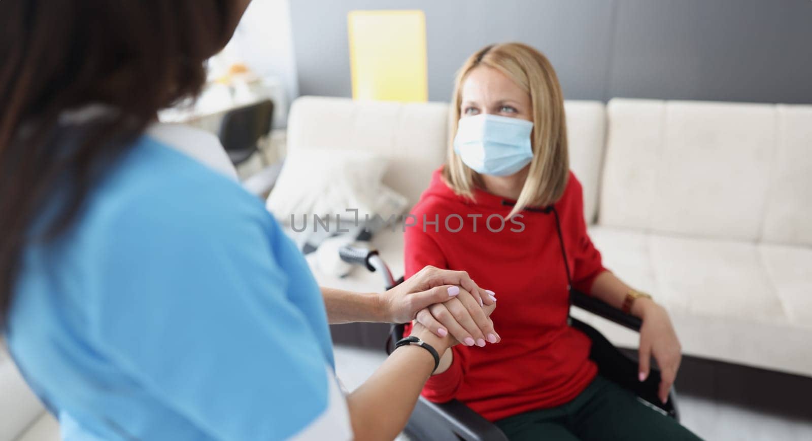 Doctor holding hand of disabled woman in wheelchair in protective medical mask by kuprevich