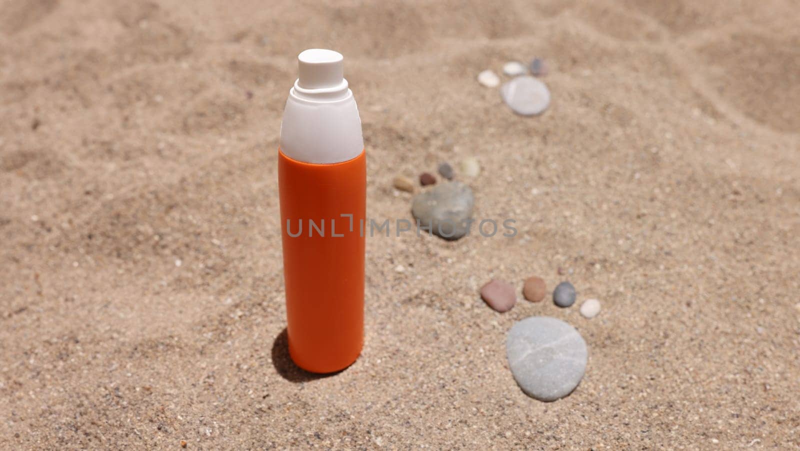 Orange jar of sunscreen standing on sand near stone footprints closeup by kuprevich