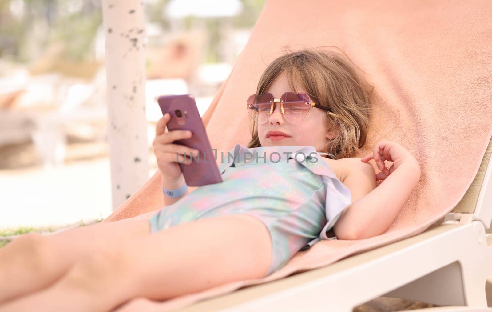 Little girl in sunglasses and swimsuit lying on lounger with mobile phone in her hands by kuprevich