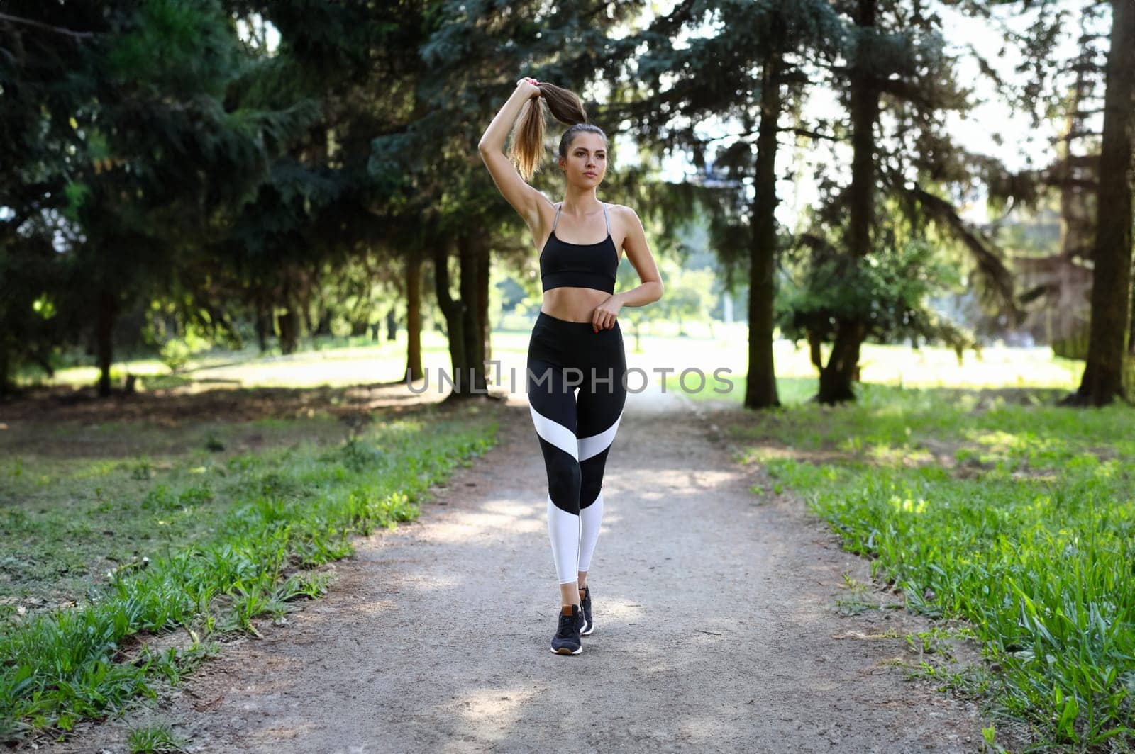 Slender girl in sportswear straightens her hair in park. Sports training at same time leads to self-discipline. Athlete circulatory system works more efficiently. Beautiful girl walks in park.