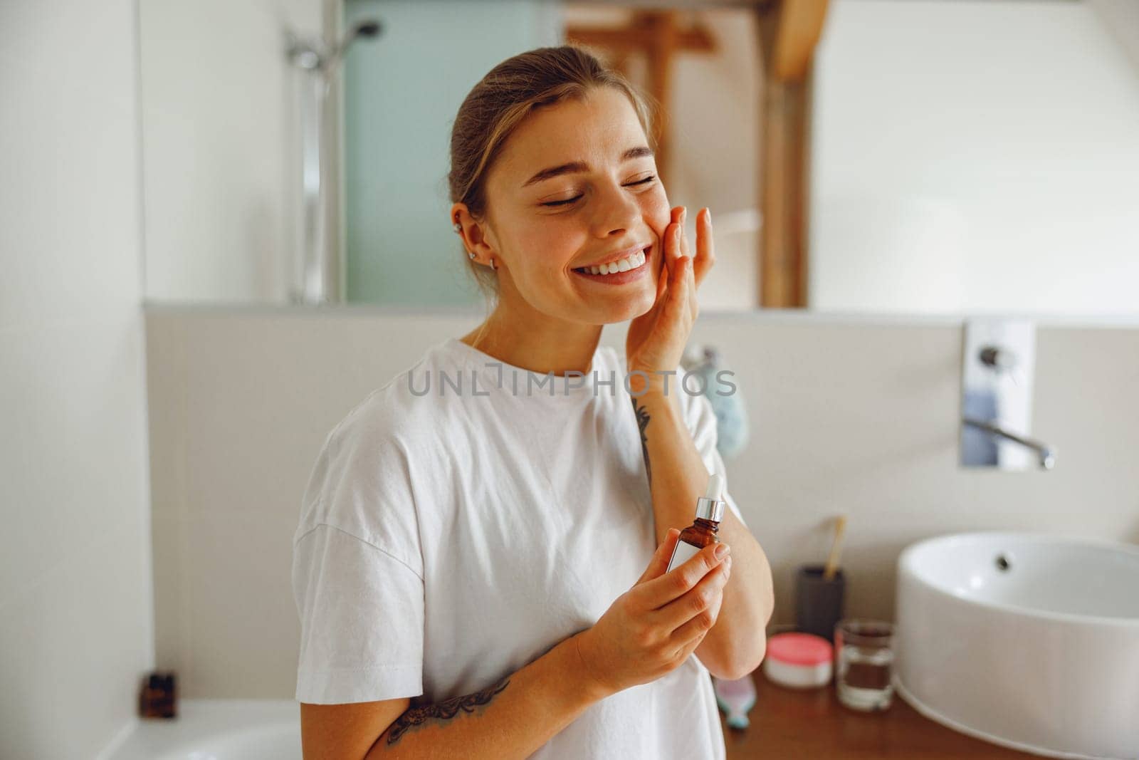 Beautiful young woman applying cosmetic serum onto her face in bathroom. Facial Skincare