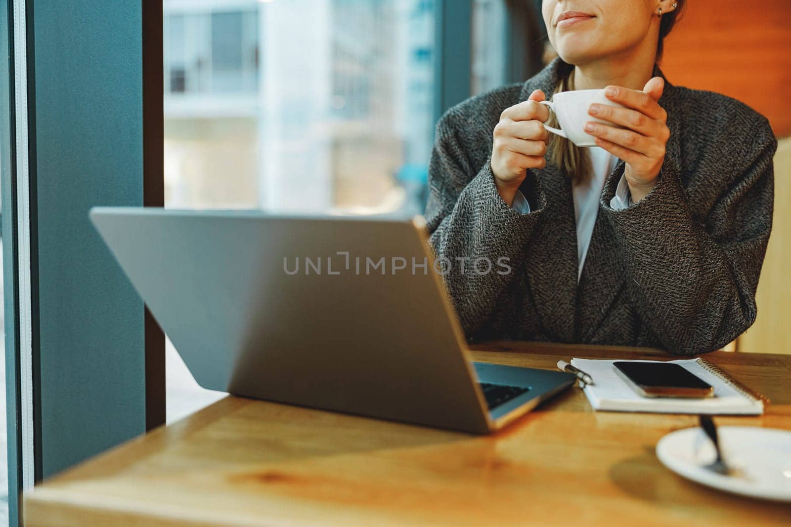 Close up of female entrepreneur is drinking coffee in coworking while working on laptop near window