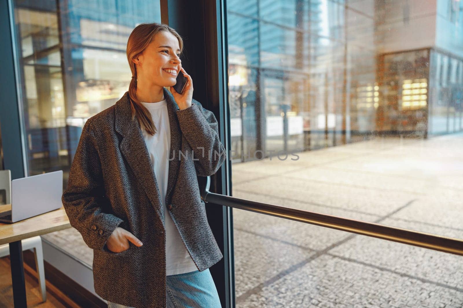 Smiling businesswoman is talking phone with colleagues while standing in cafe near window