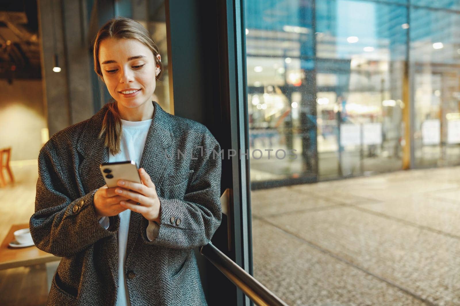 Young female entrepreneur is use phone while standing in cafe near window. Distance work concept by Yaroslav_astakhov