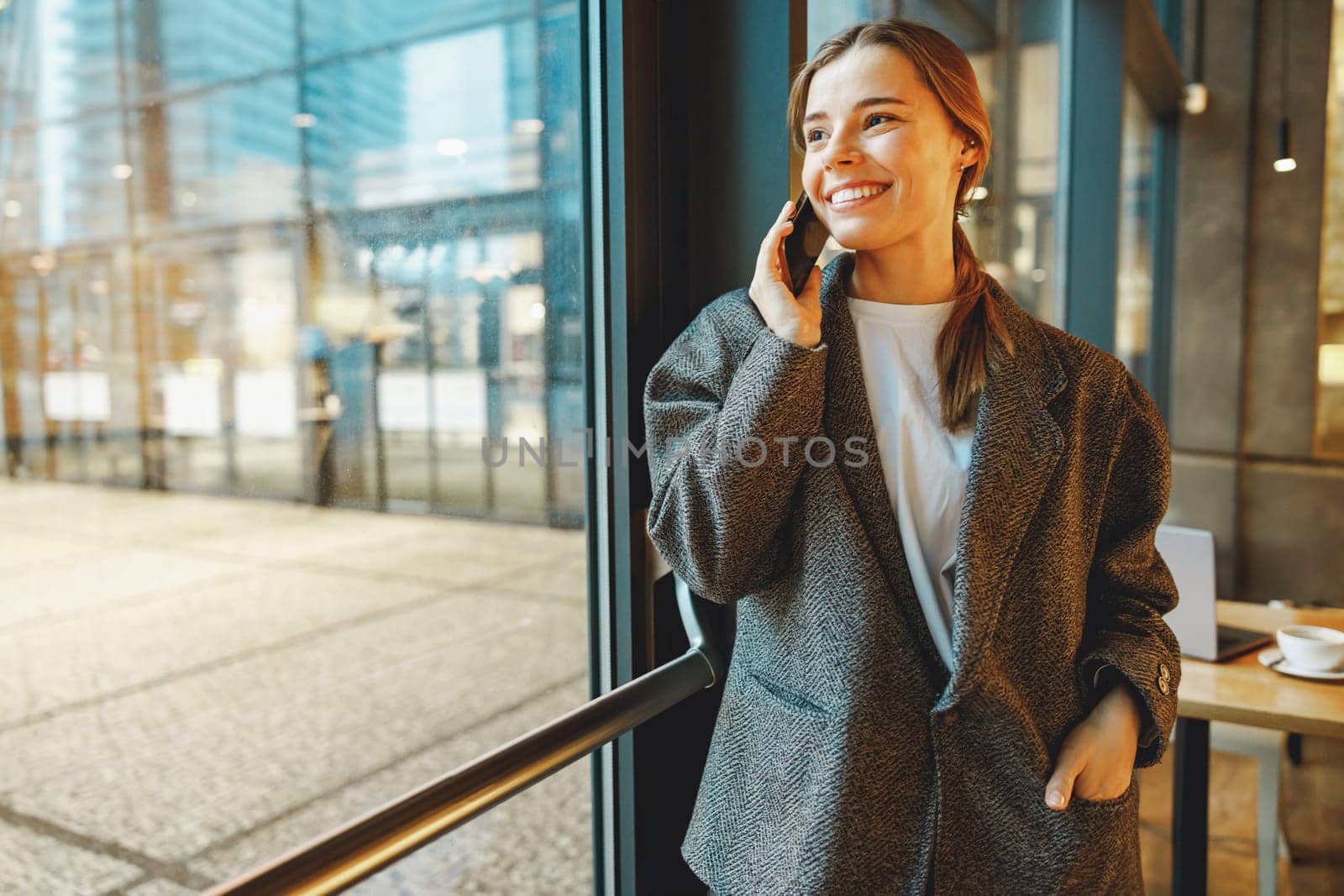 Stylish young female freelancer is talking phone with client while standing in coworking near window