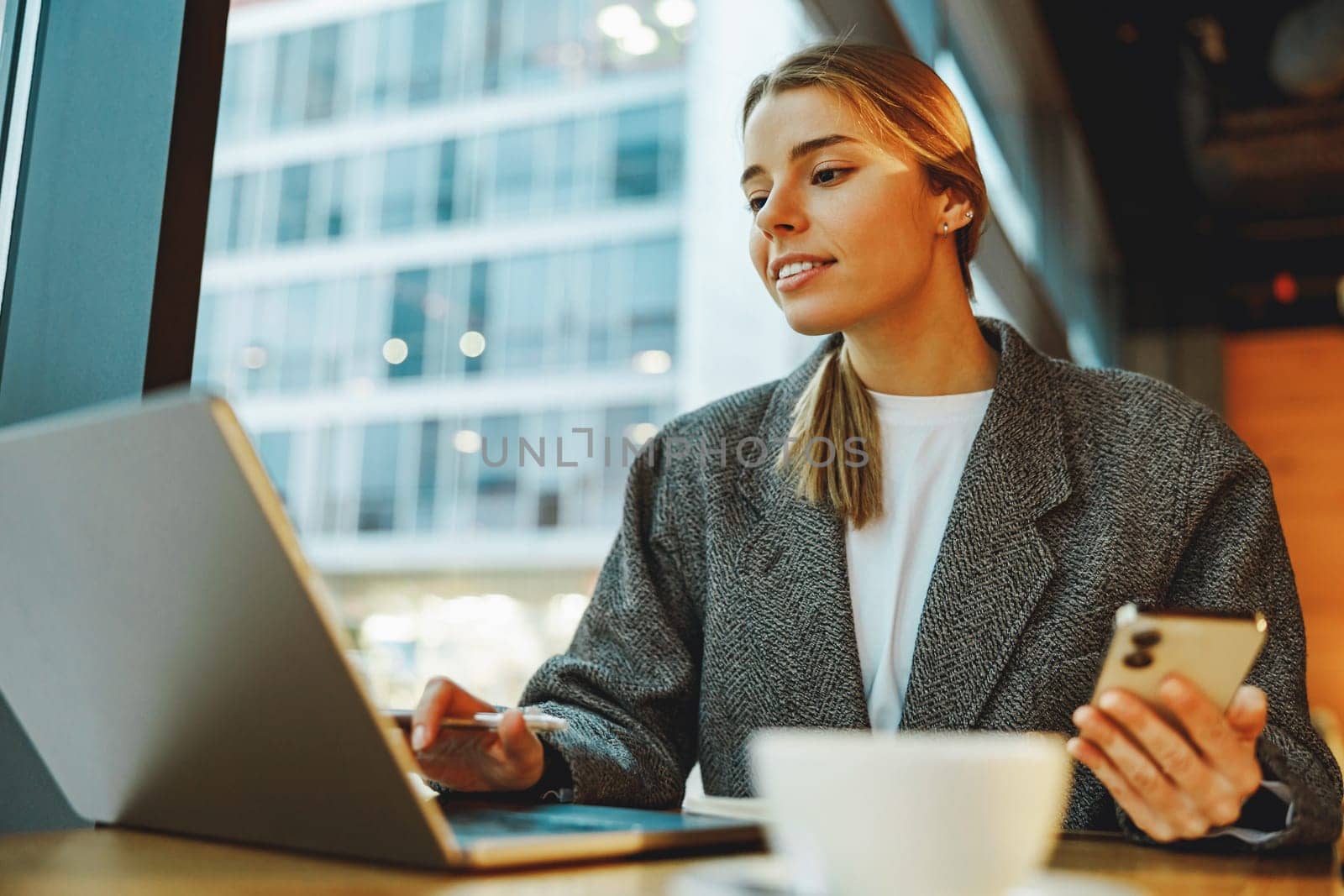 Woman manager reading message on phone and work on laptop in cafe. Distance work concept