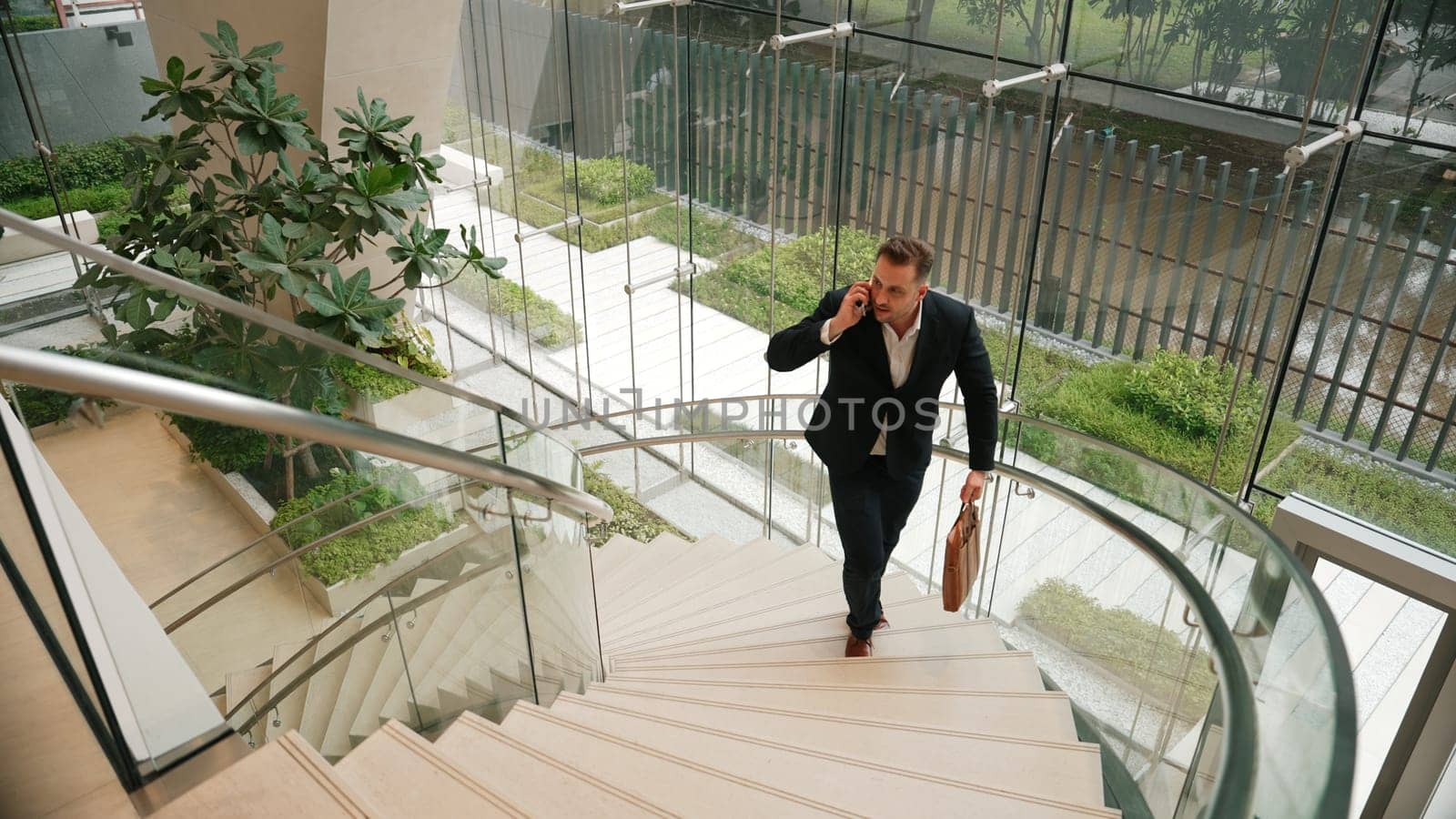 Smart businessman walking up spiral stair case and calling manager by using smart phone. Top view of manager talking marketing team by using telephone while going up stair at modern building. Urbane.