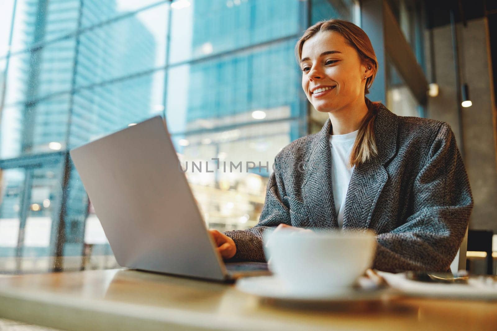 Smiling female entrepreneur is drinking coffee in coworking while working on laptop near window by Yaroslav_astakhov