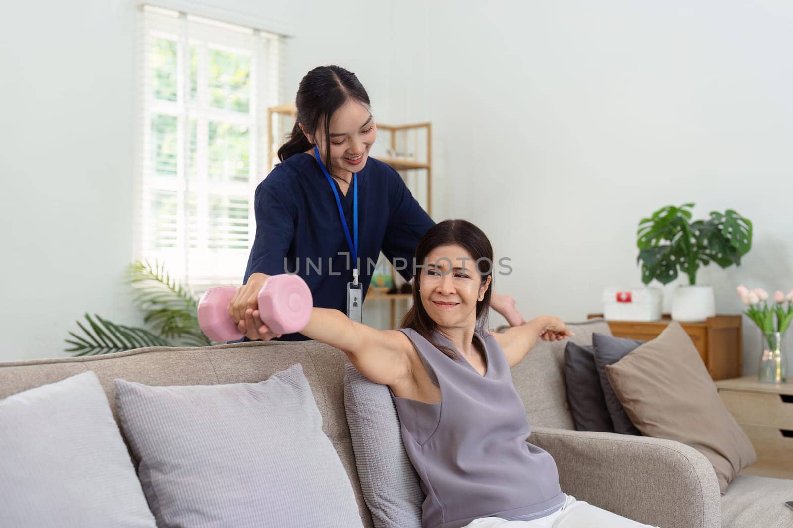 Asian older woman doing physiotherapist with support from nurse or caregiver. Senior elderly woman using dumbbells workout exercise for patient with caregiver in nursing care at home by itchaznong