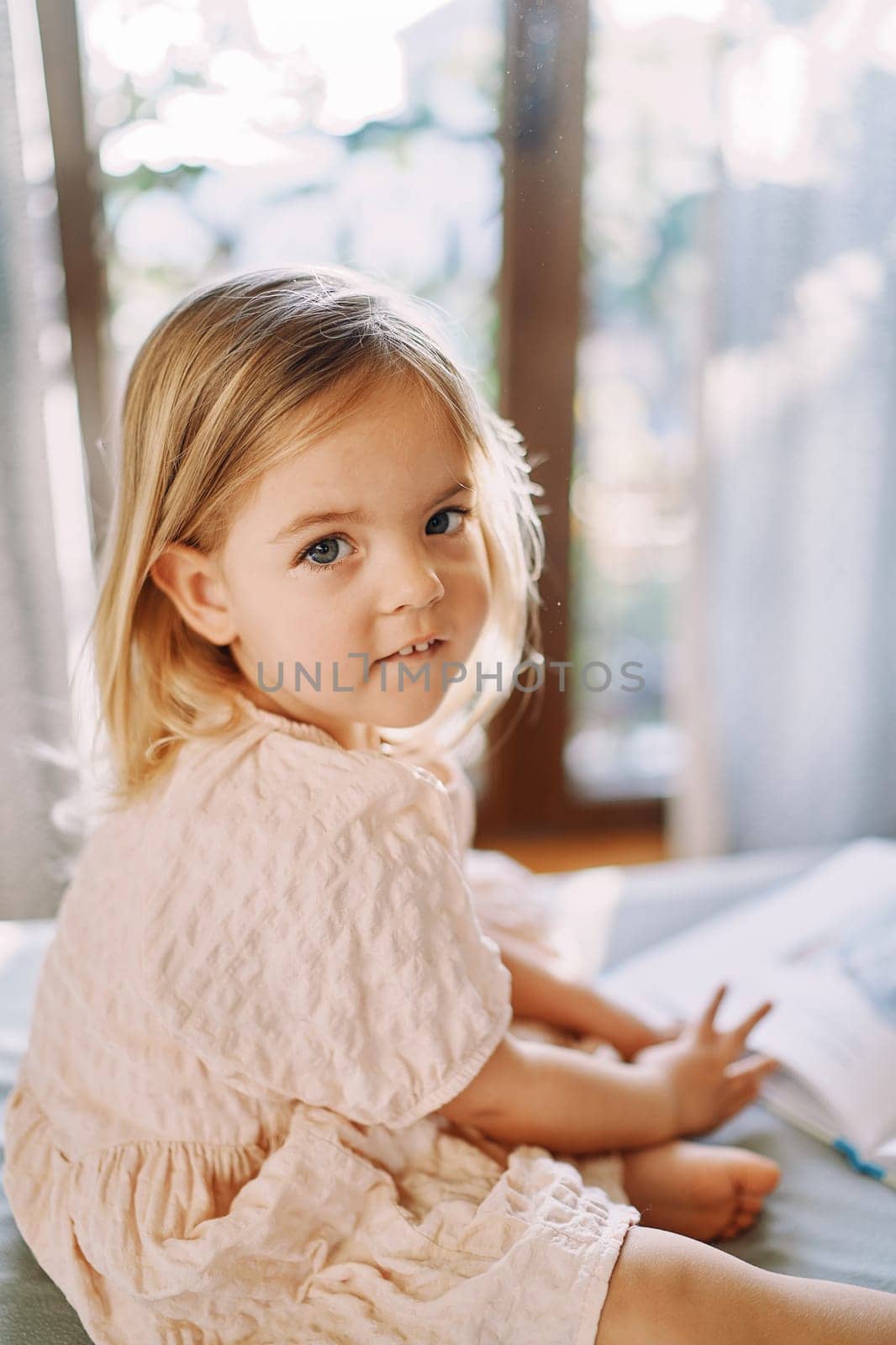 Little girl sits on the bed with a book and looks to the side. Side view. High quality photo