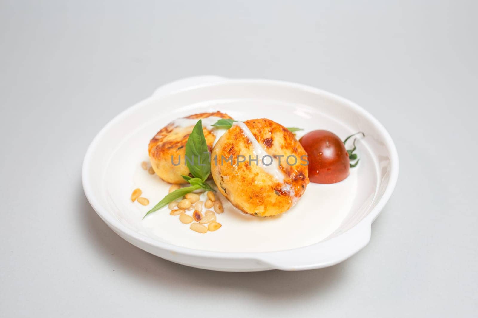 Two round fried cheese pancakes with sour cream and cherry tomatoes on a white plate on a white background. Isolated on a white background.