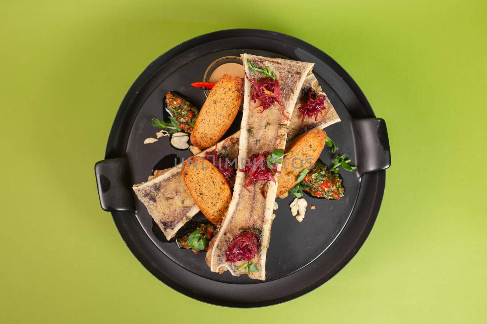 Top view of beef bone marrow on a black plate with toast and greens on a green background. The delicacy is on the menu. by Pukhovskiy