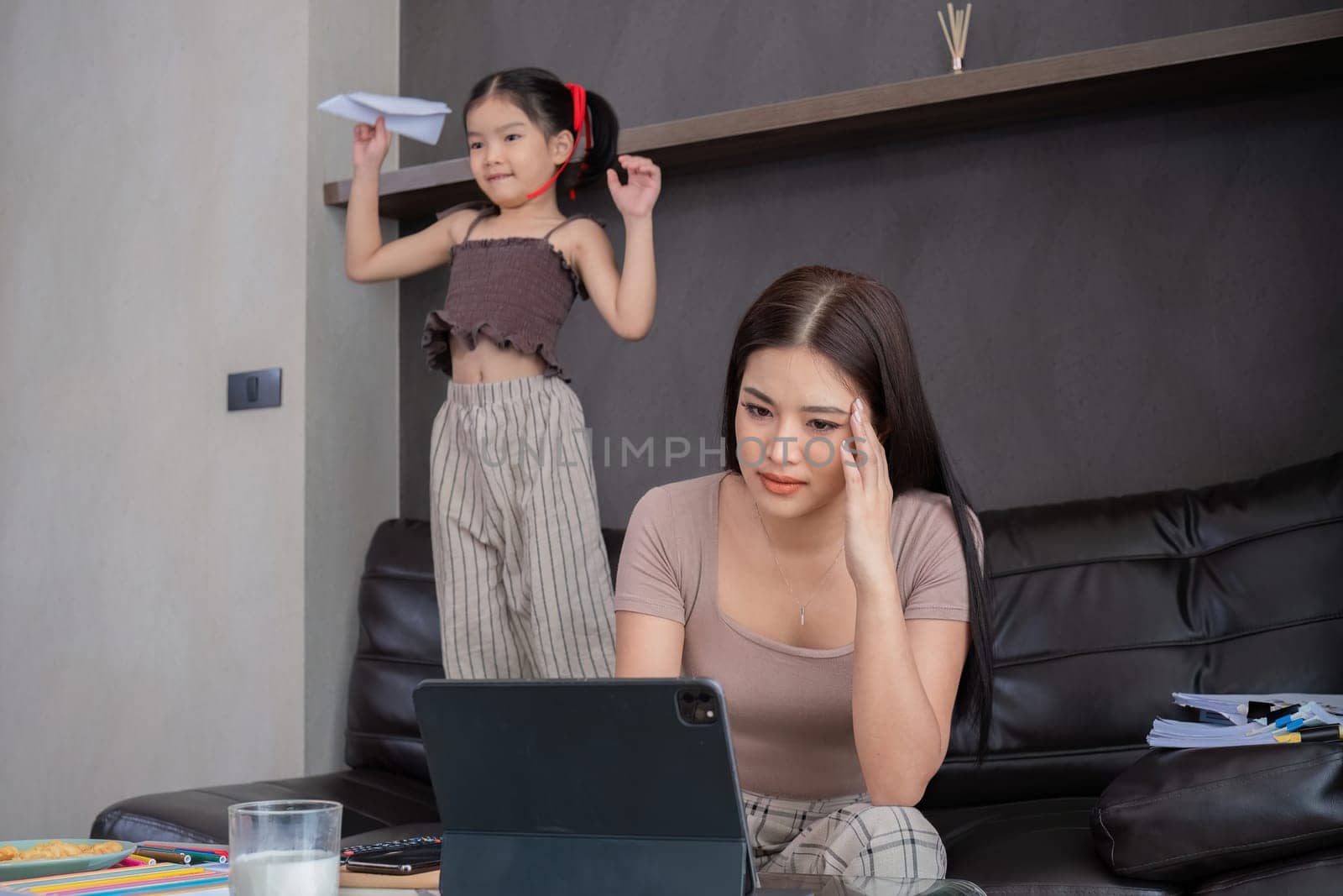 Stressed business mother sitting from home, taking notes and looking after her laptop with her cute daughter playing next to her..