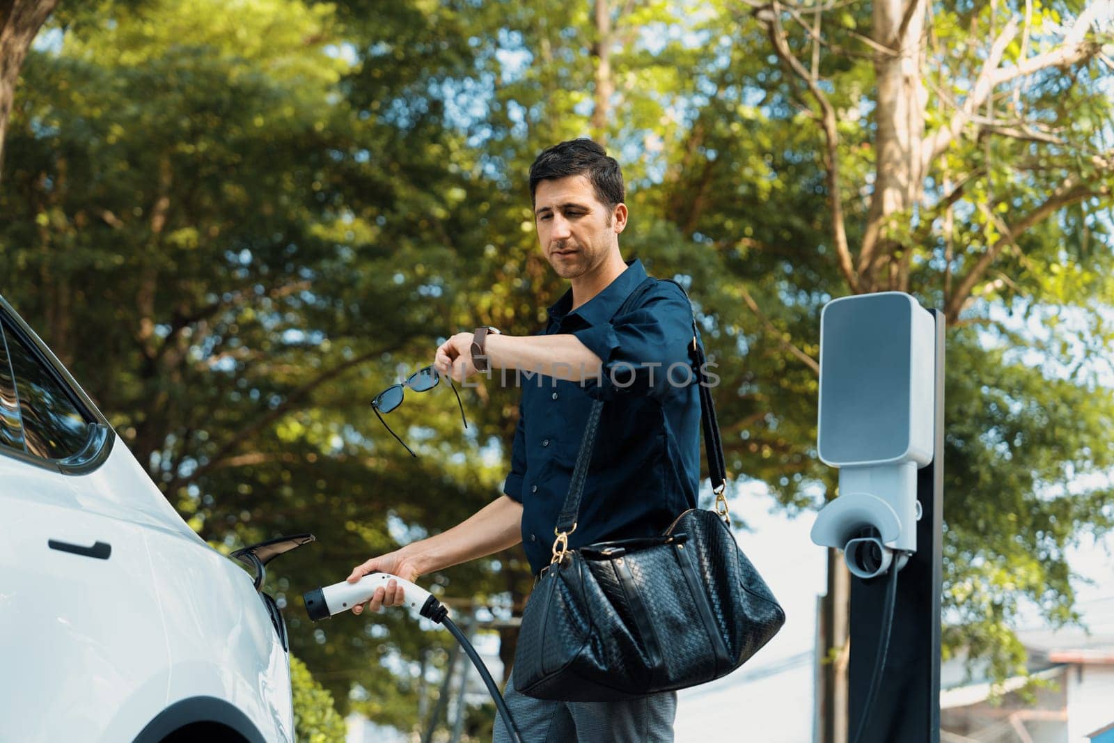 Young man recharge electric car's battery from charging station in outdoor green city park in springtime. Rechargeable EV car for sustainable environmental friendly urban travel lifestyle. Expedient