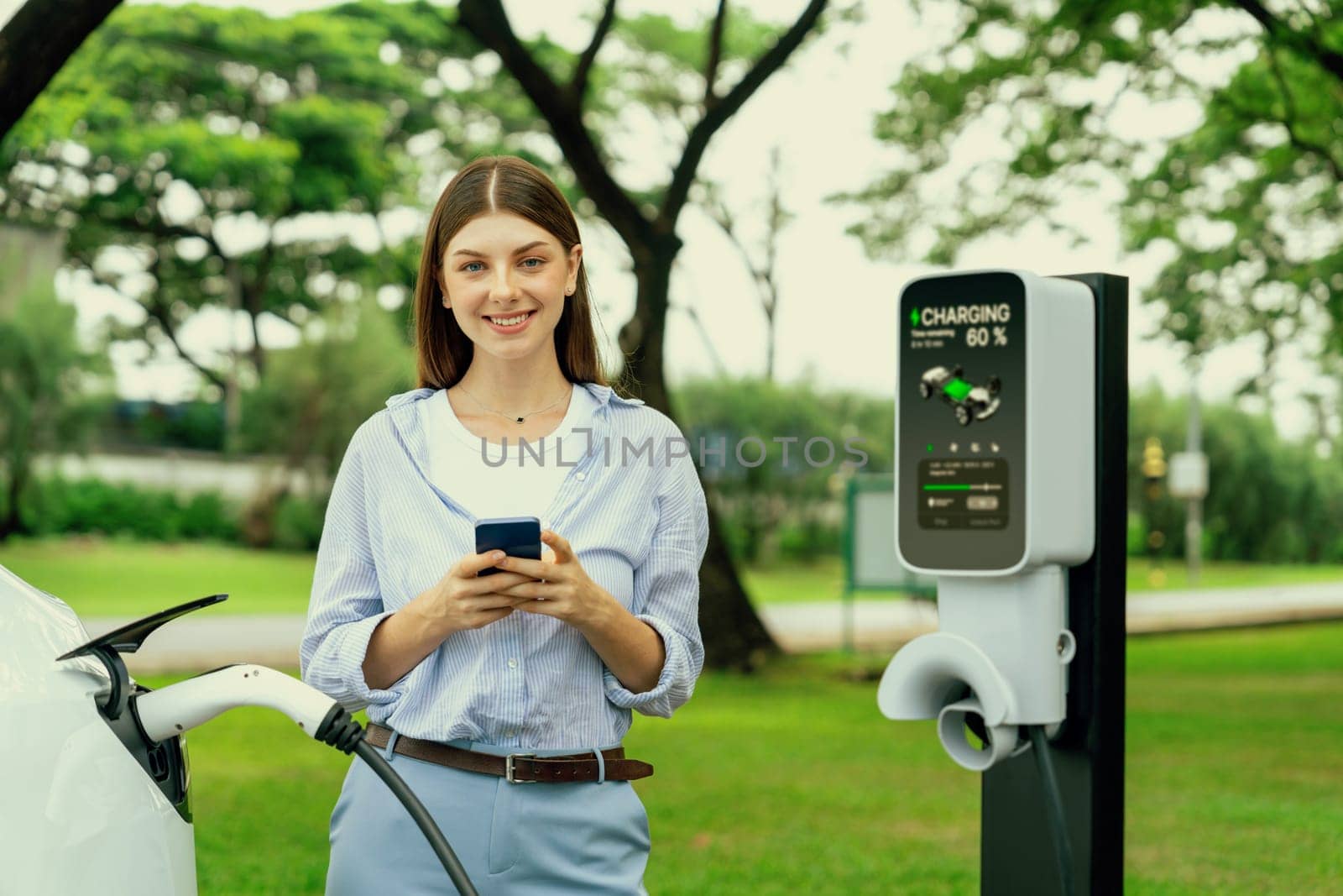 Young woman recharge EV electric vehicle battery from EV charging station and using smartphone online banking to pay for electricity in city park. Eco friendly vehicle travel with EV car. Exalt