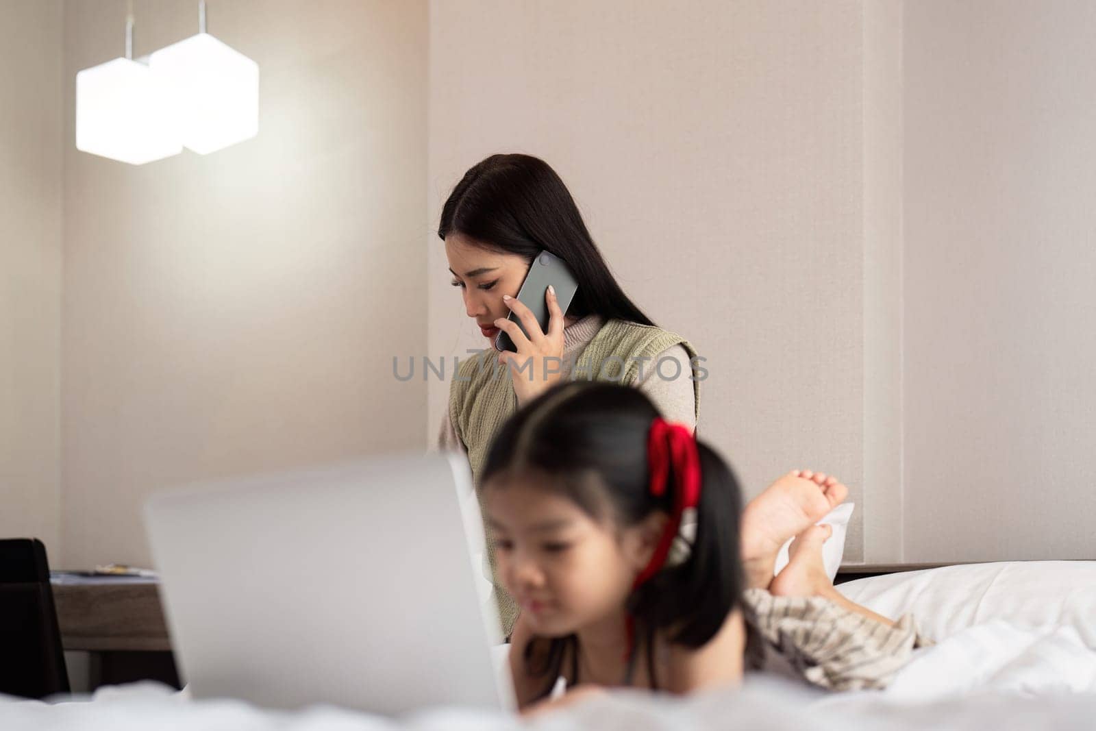 Asian woman in casual outfit is talking on the mobile phone and working on laptop at home with her daughter.