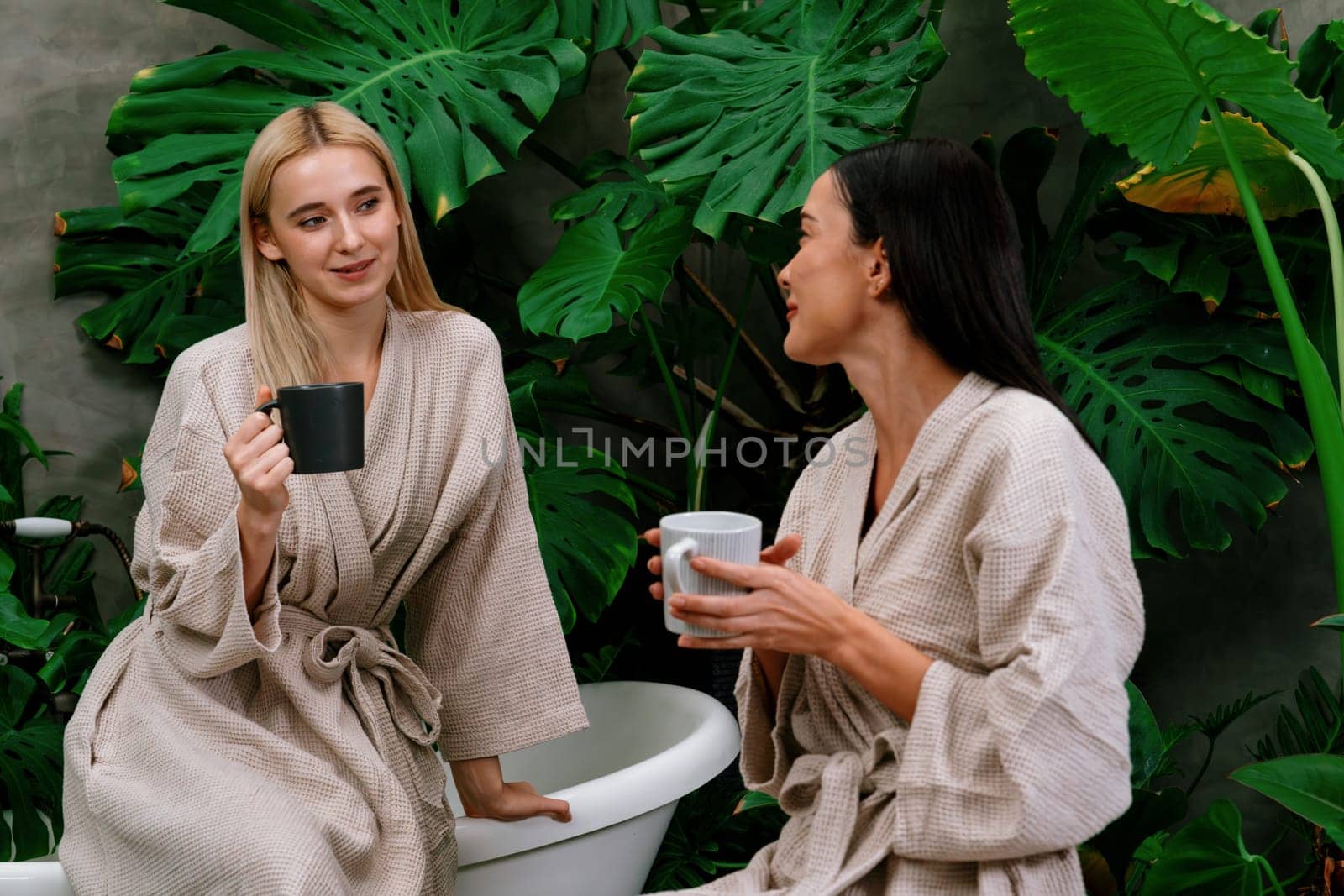 Tropical and exotic spa garden with bathtub in modern hotel or resort with young two women in bathrobe drink coffee, enjoy leisure and wellness lifestyle surround by lush greenery foliage. Blithe