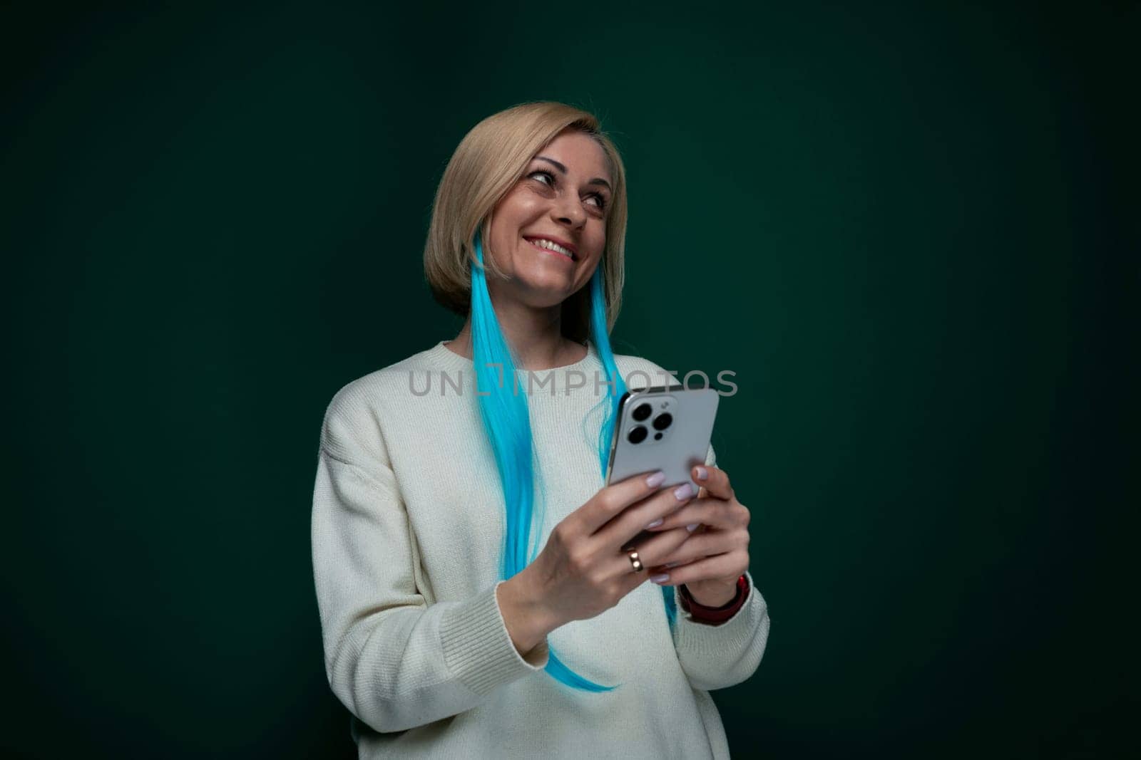 A woman with vibrant blue hair is seen holding a cell phone in her hand. She appears focused on the devices screen, possibly texting or browsing. The background is simple and nondescript.