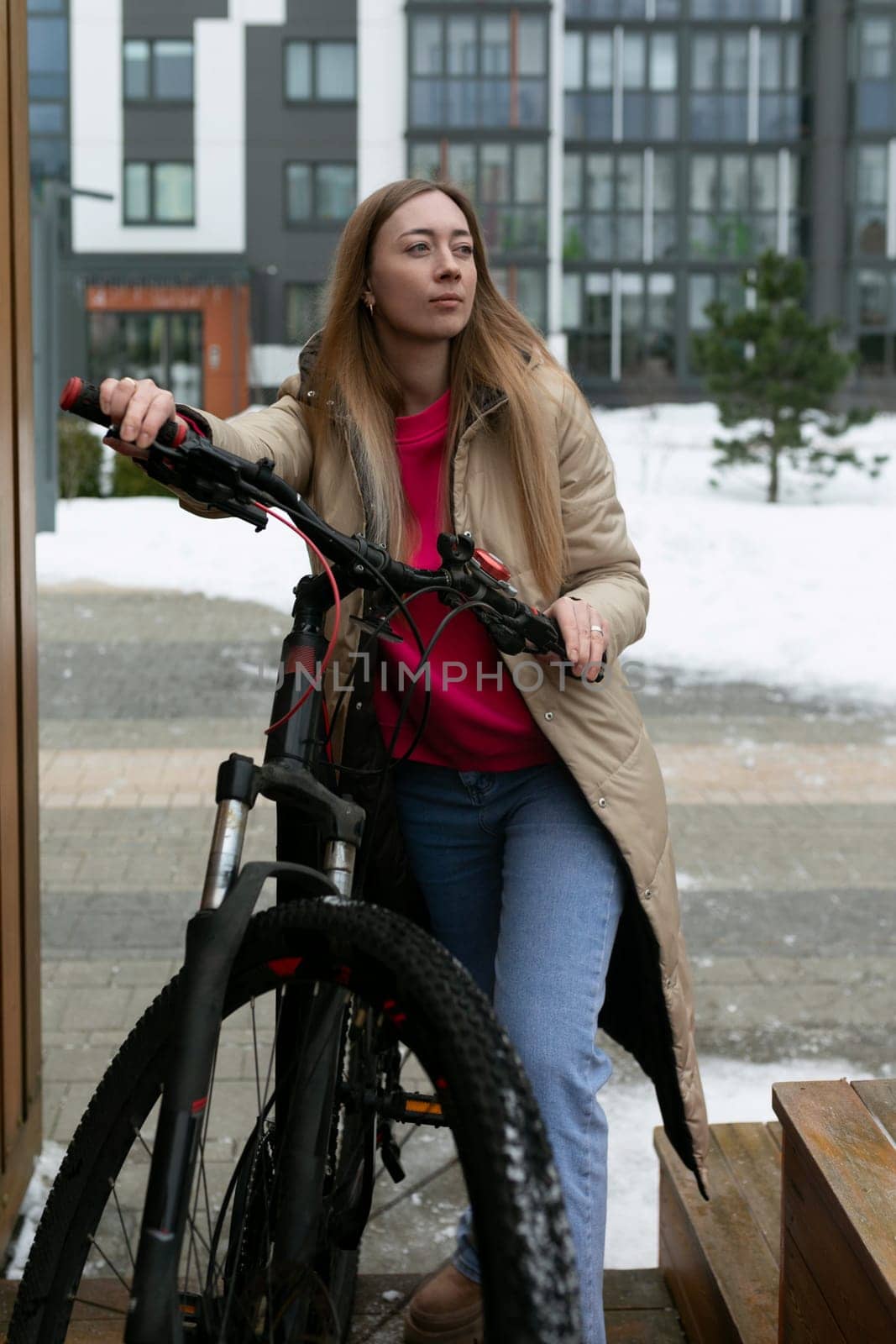 A woman in casual attire sitting on a wooden bench next to a black bicycle. The woman appears relaxed and is looking ahead. The bike is parked beside her on the pavement.