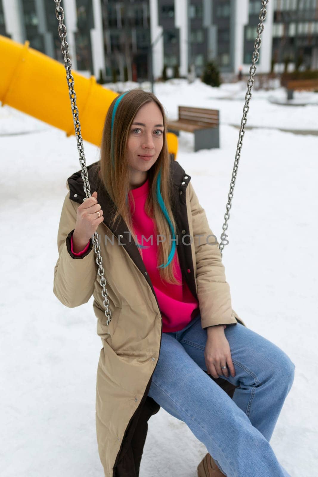 A woman is seated on a swing, swinging gently back and forth, in a snowy outdoor setting. Snow covers the ground and tree branches, creating a winter wonderland scene.
