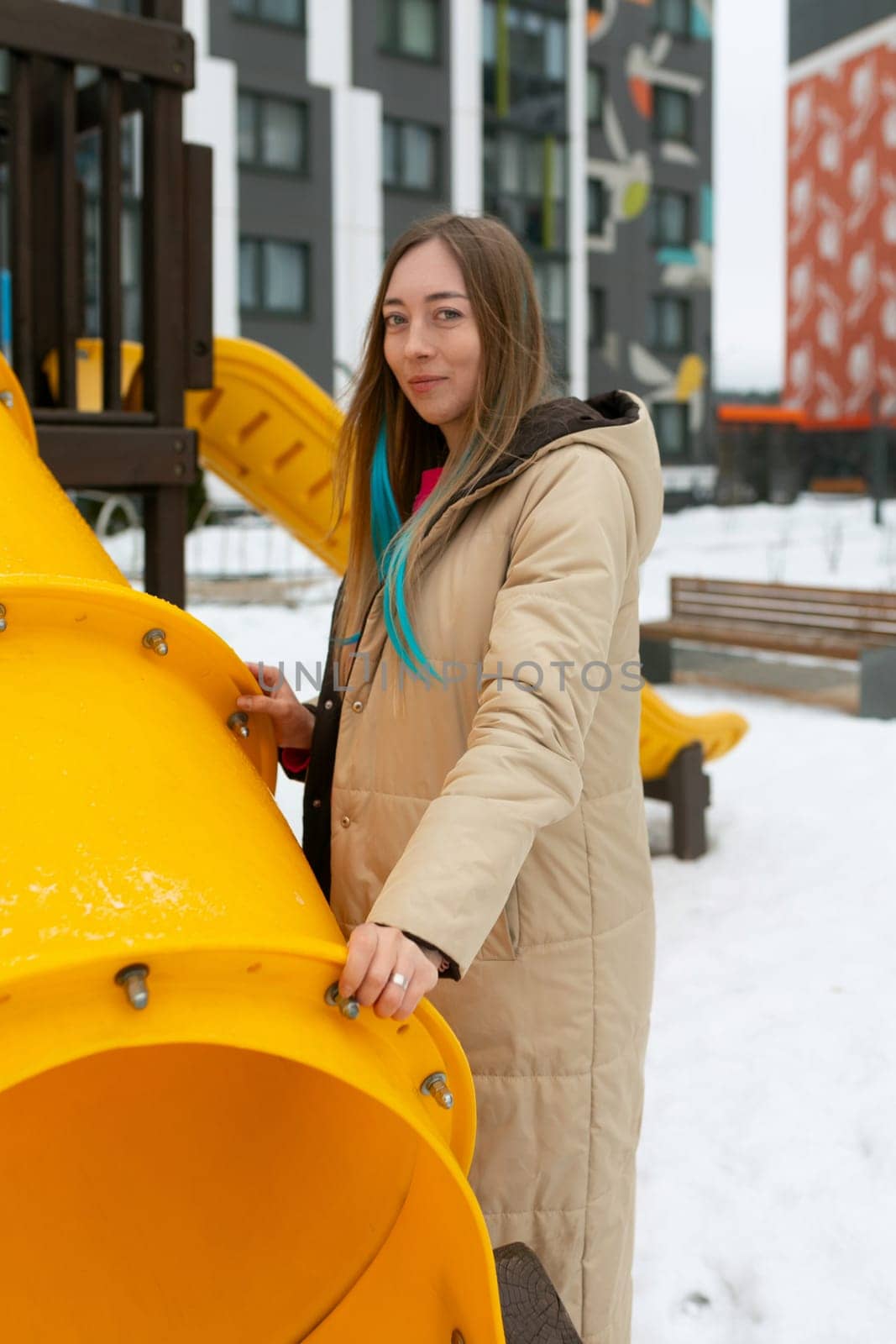 A woman is standing next to a bright yellow playground structure, looking towards the camera. The playground equipment features slides, stairs, and platforms. The scene is set in a sunny park with green grass and clear blue skies.