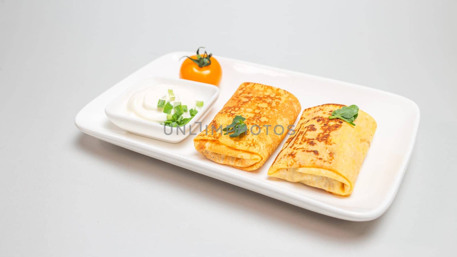 Two folded stuffed savory crepes with sour cream and chives garnish on a white plate on a white background.