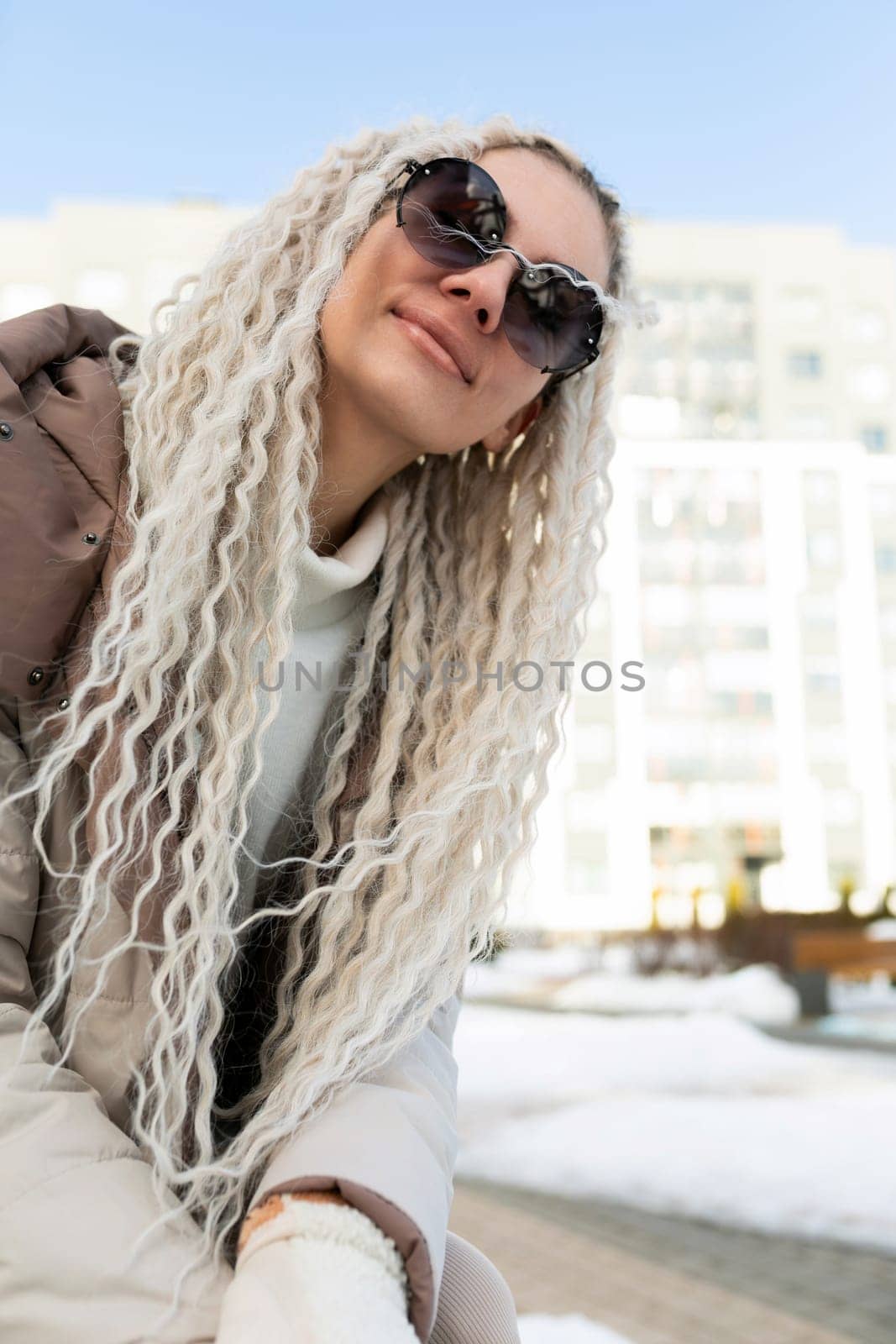A woman with long blonde hair is wearing stylish sunglasses. She looks confident and fashionable as she stands in the sunlight.
