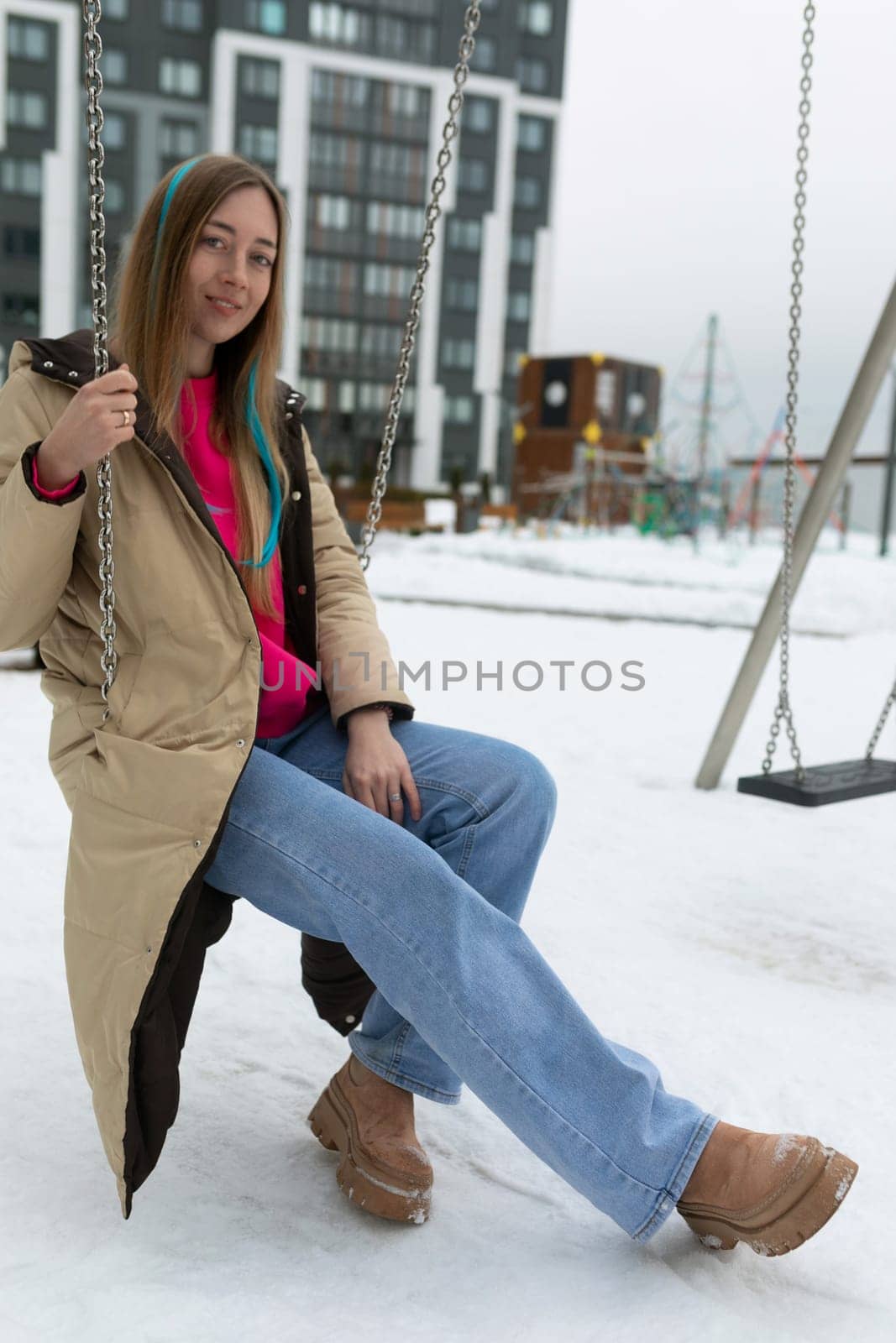 A woman wearing winter clothing sits on a swing covered in snow. The snowflakes are gently falling around her, creating a serene winter scene.