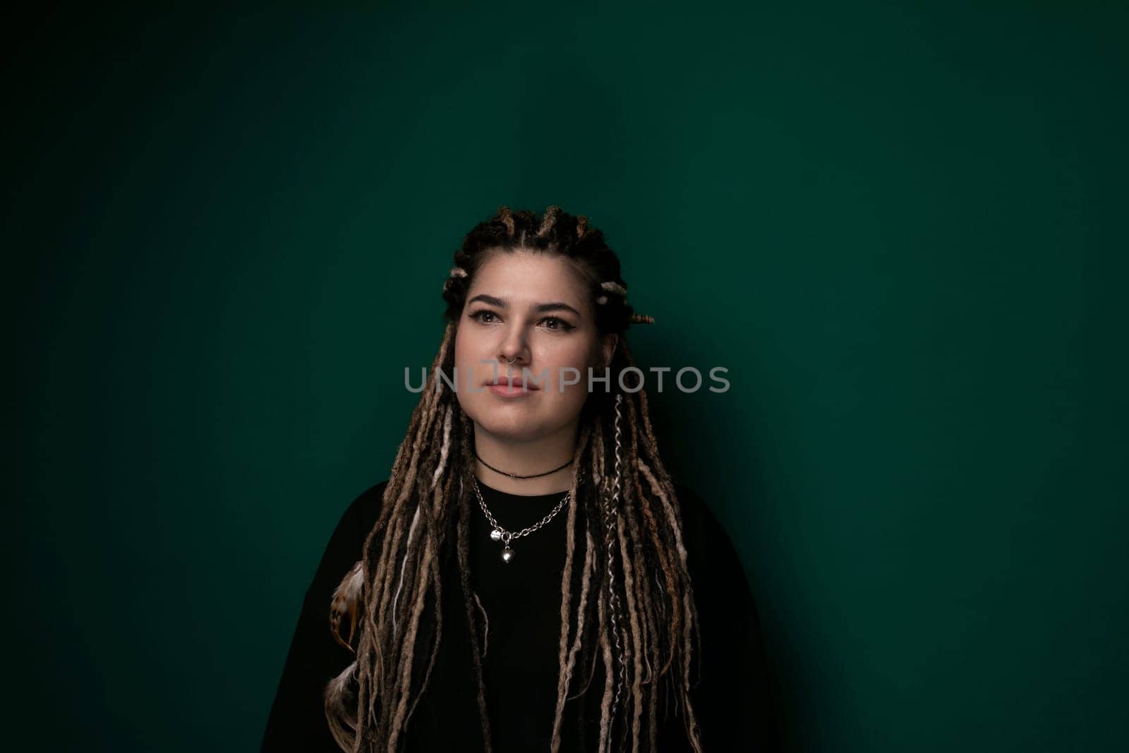 A woman with dreadlocks is standing confidently in front of a vibrant green wall, showcasing her unique hairstyle. The bright green color of the wall contrasts with her dark hair, creating a striking visual.