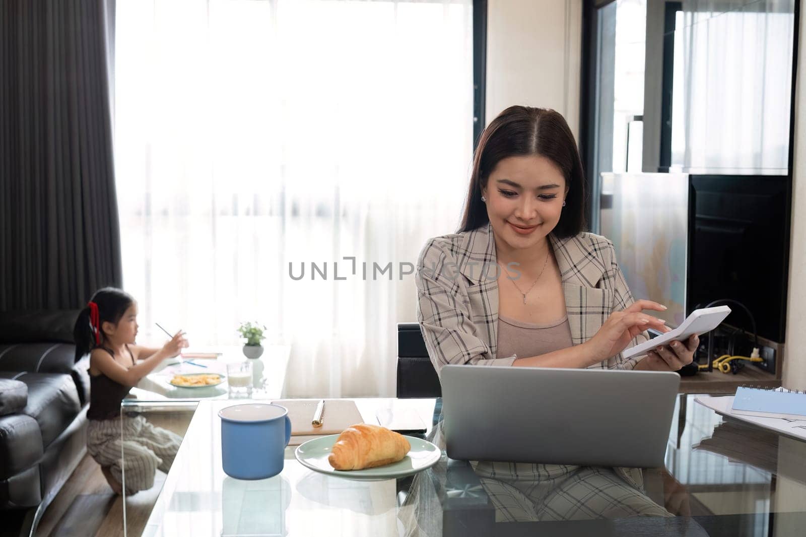 A business mother works from home, doing math, taking notes and tending to her laptop with her adorable daughter intent on drawing behind her..