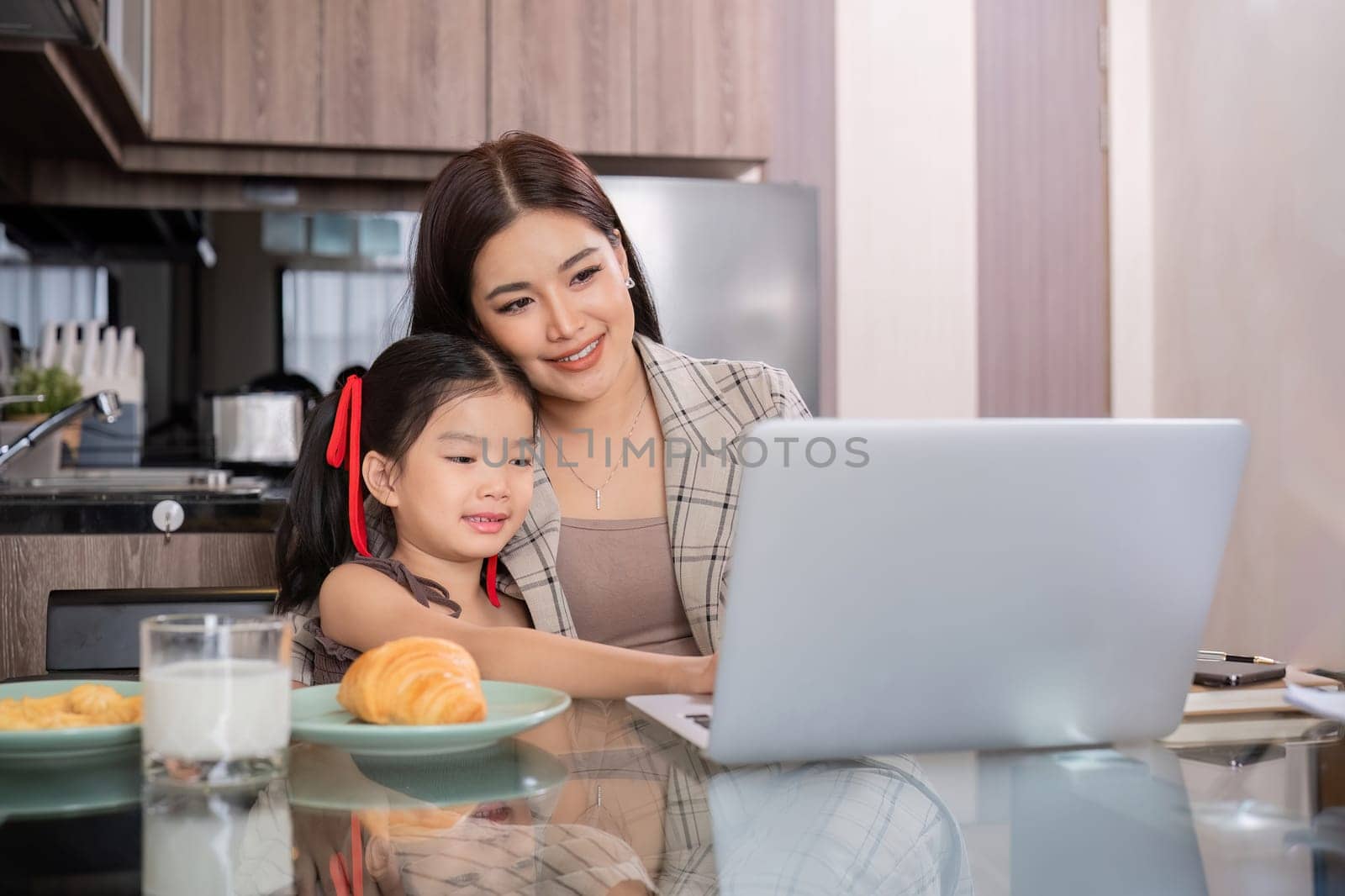 a single mother, sits at home working on a laptop with her daughter beside her watching and encouraging her. by wichayada