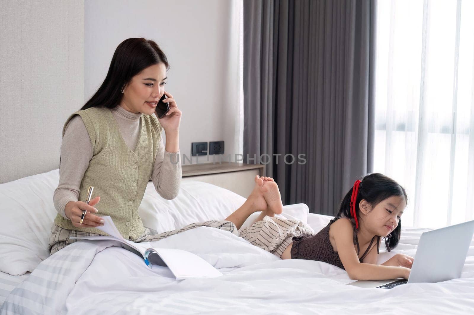 Single mother, businessman and cute daughter in bedroom A young woman is working online on a laptop, using the phone to talk about work from home, with her daughter lying beside her giving support..