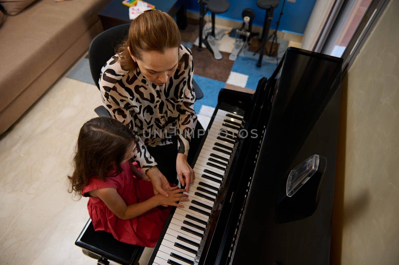 View from above of a Caucasian pleasant mature female music teacher, musician pianist teaching piano, explaining to a little kid girl student the correct position of fingers on the piano keys.