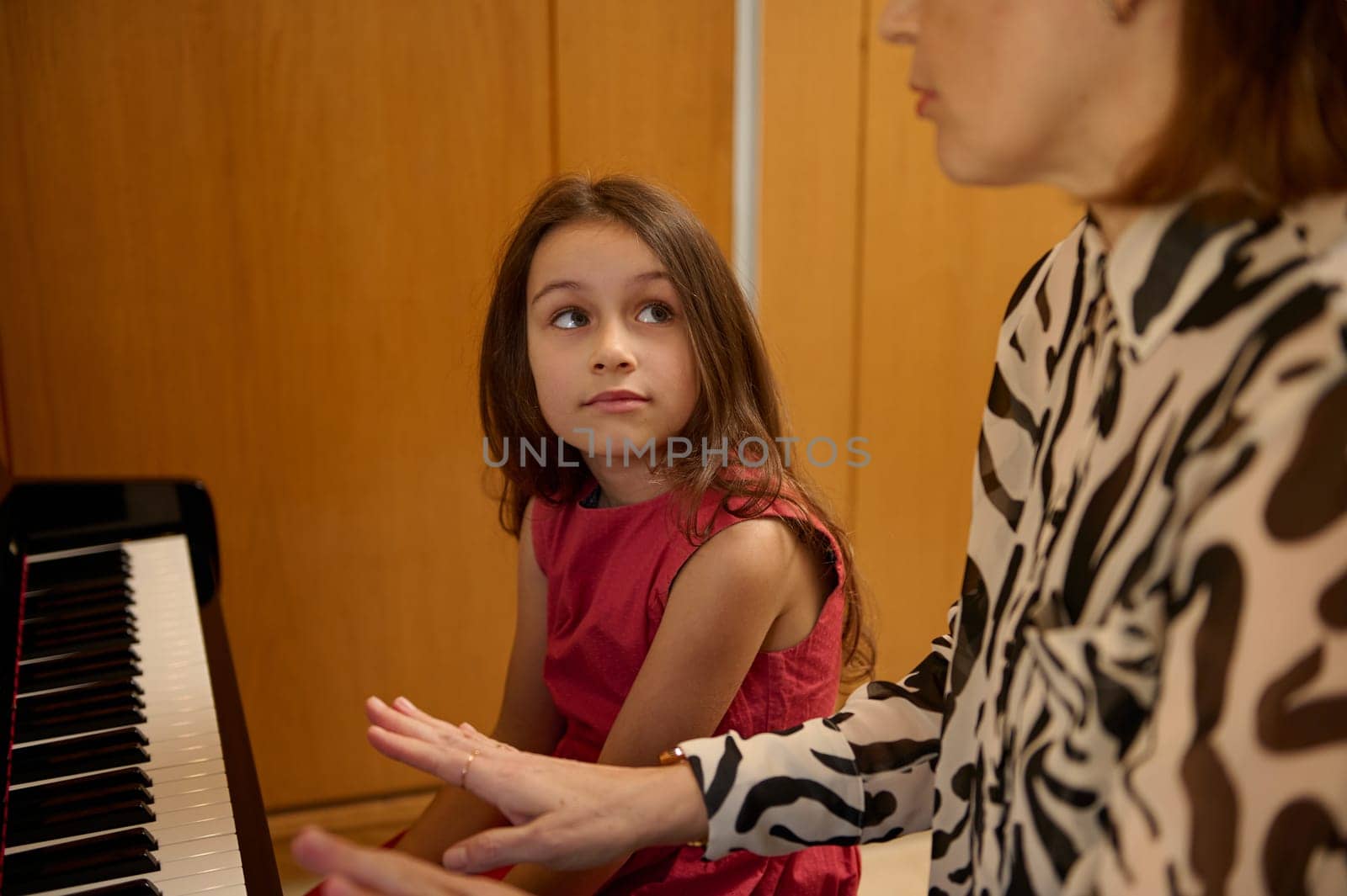 Authentic portrait of beautiful Caucasian elementary age schoolkid girl in stylish red dress, smiling looking at her music teacher, sitting together at piano during individual music lesson indoors