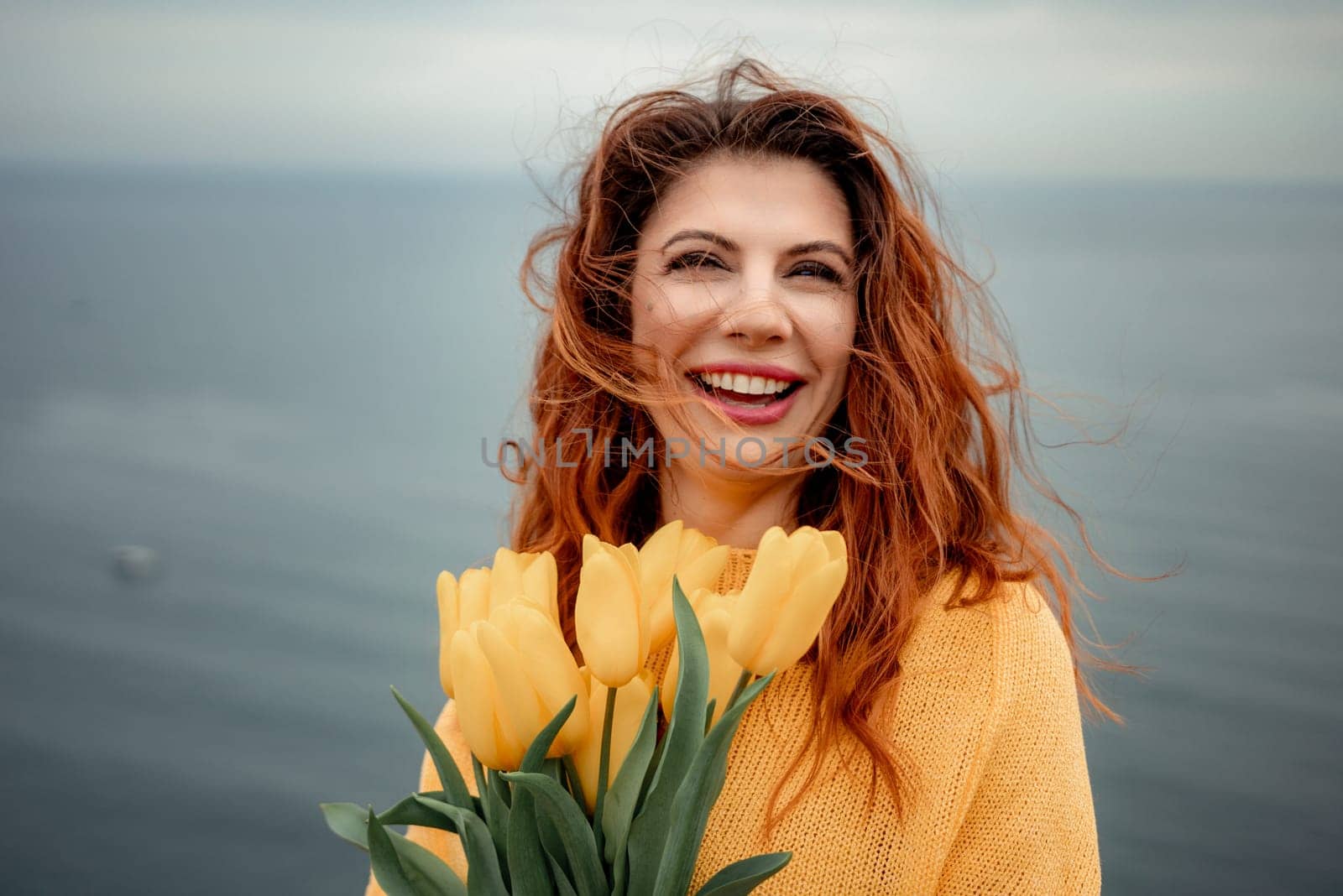 Portrait of a happy woman with hair flying in the wind against the backdrop of mountains and sea. Holding a bouquet of yellow tulips in her hands, wearing a yellow sweater by Matiunina