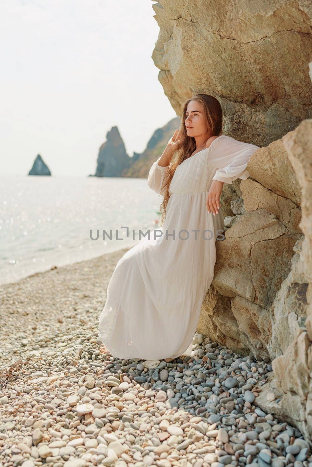 woman white dress standing on a beach, looking out at the ocean. She is lost in thought, with her hand on her hip. Concept of calm and introspection