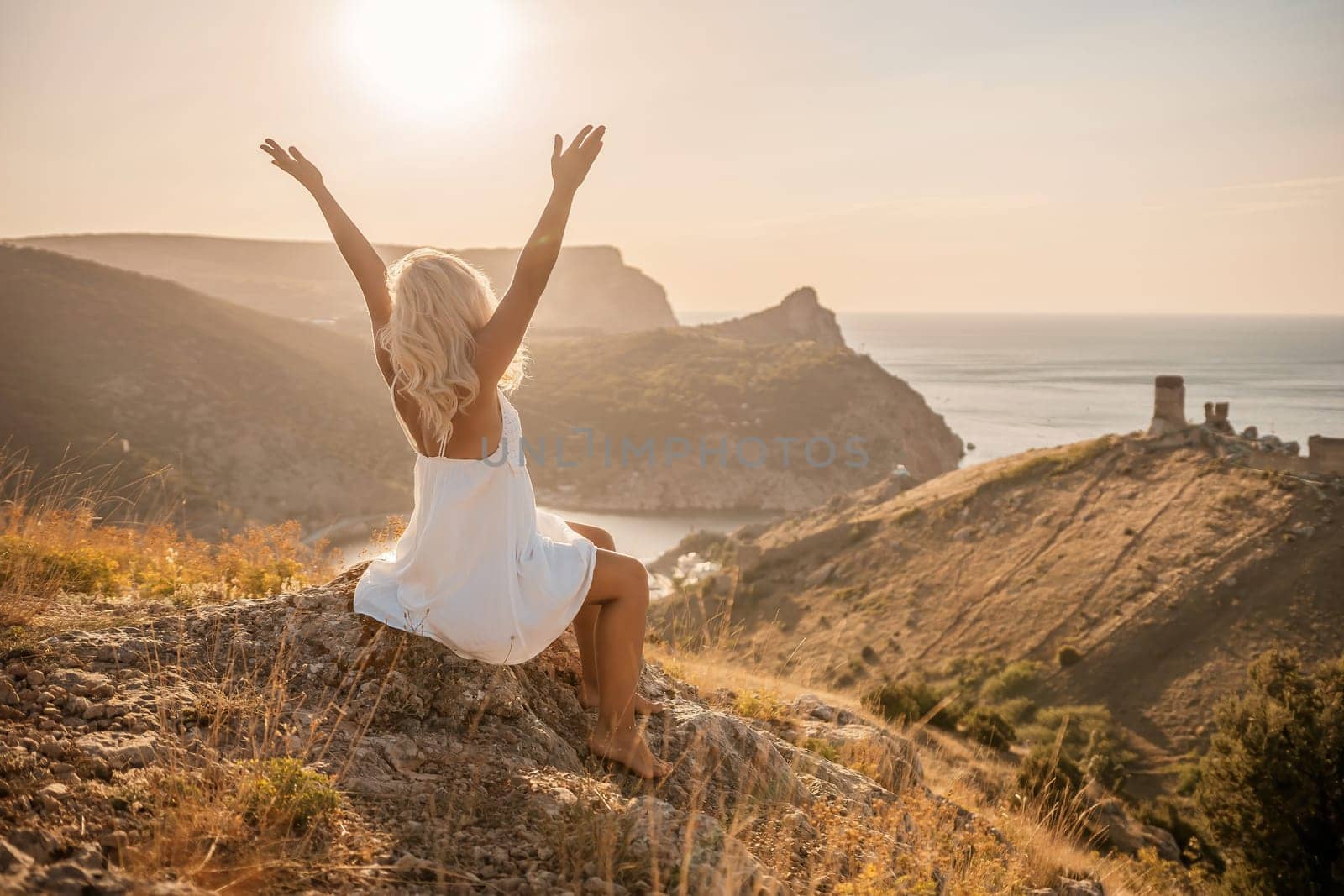Happy woman is sitting on a hillside, wearing a white dress. She is surrounded by a beautiful landscape, with a body of sea in the background. Concept of peace and happiness