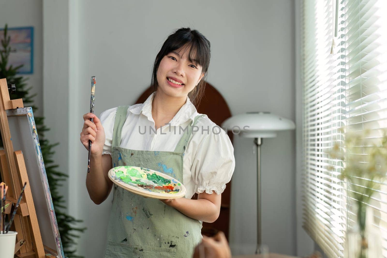 A young artistic girl holds a paintbrush and a paint tray and looks cheerfully at the camera..