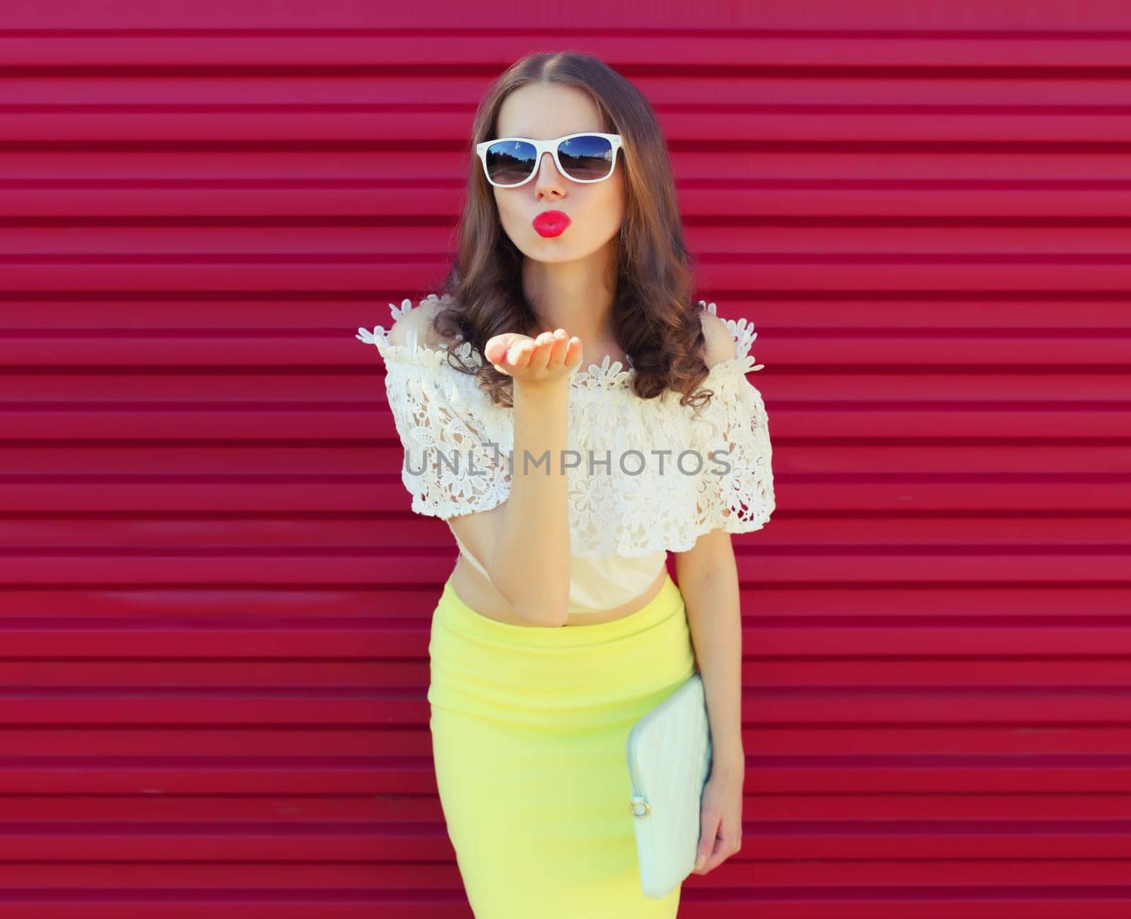 Beautiful young woman model blowing kiss posing with handbag clutch on pink background