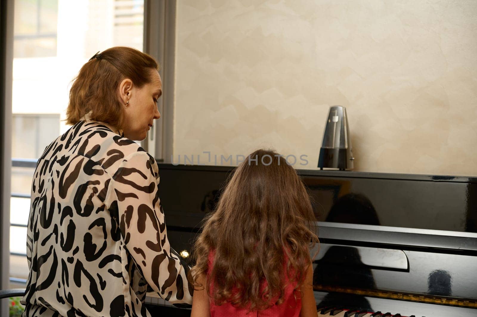 Back view young adult woman, music teacher with the pupil, sitting at vintage wooden piano at home, giving a music lesson. Mother and cute daughter playing piano together. People. Hobbies and leisure