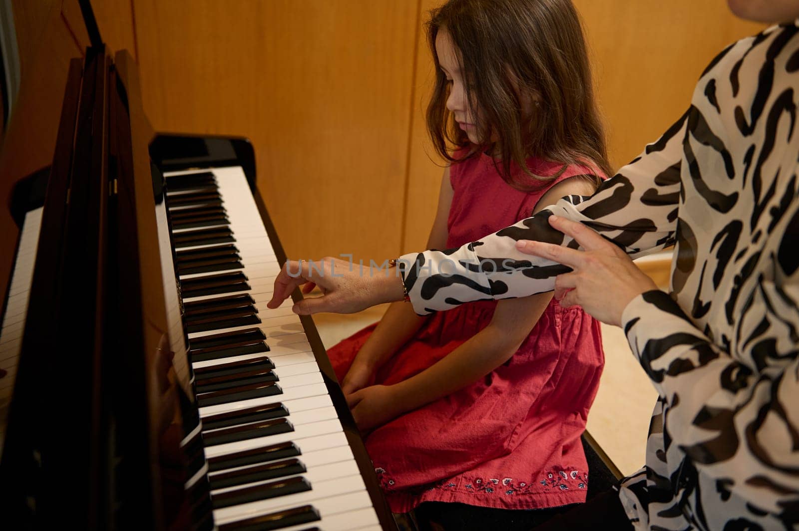 Little kid girl in elegant red dress, taking piano lesson, passionately playing the keys under her teacher's guidance, feeling the rhythm of music. Musical education and talent development in progress by artgf