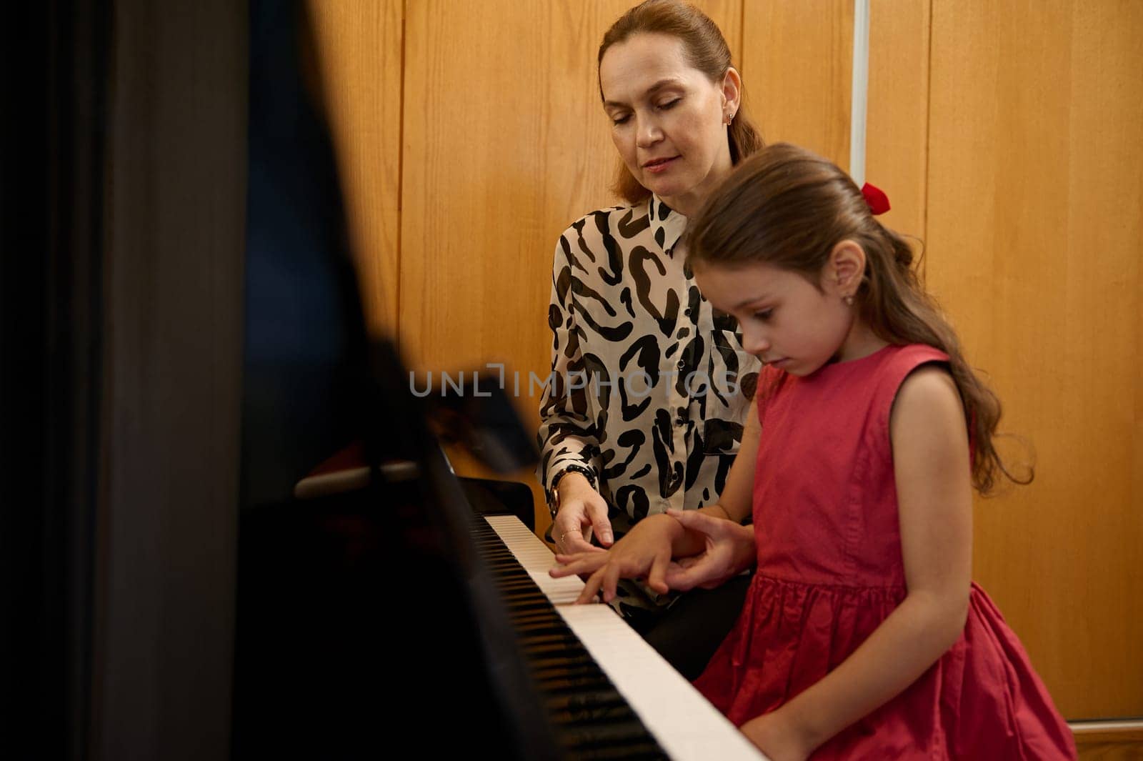 Adorable little girl in red dress, taking piano lesson, passionately playing the keys under her teacher's guidance, feeling the rhythm of melody. Musical education and talent development in progress