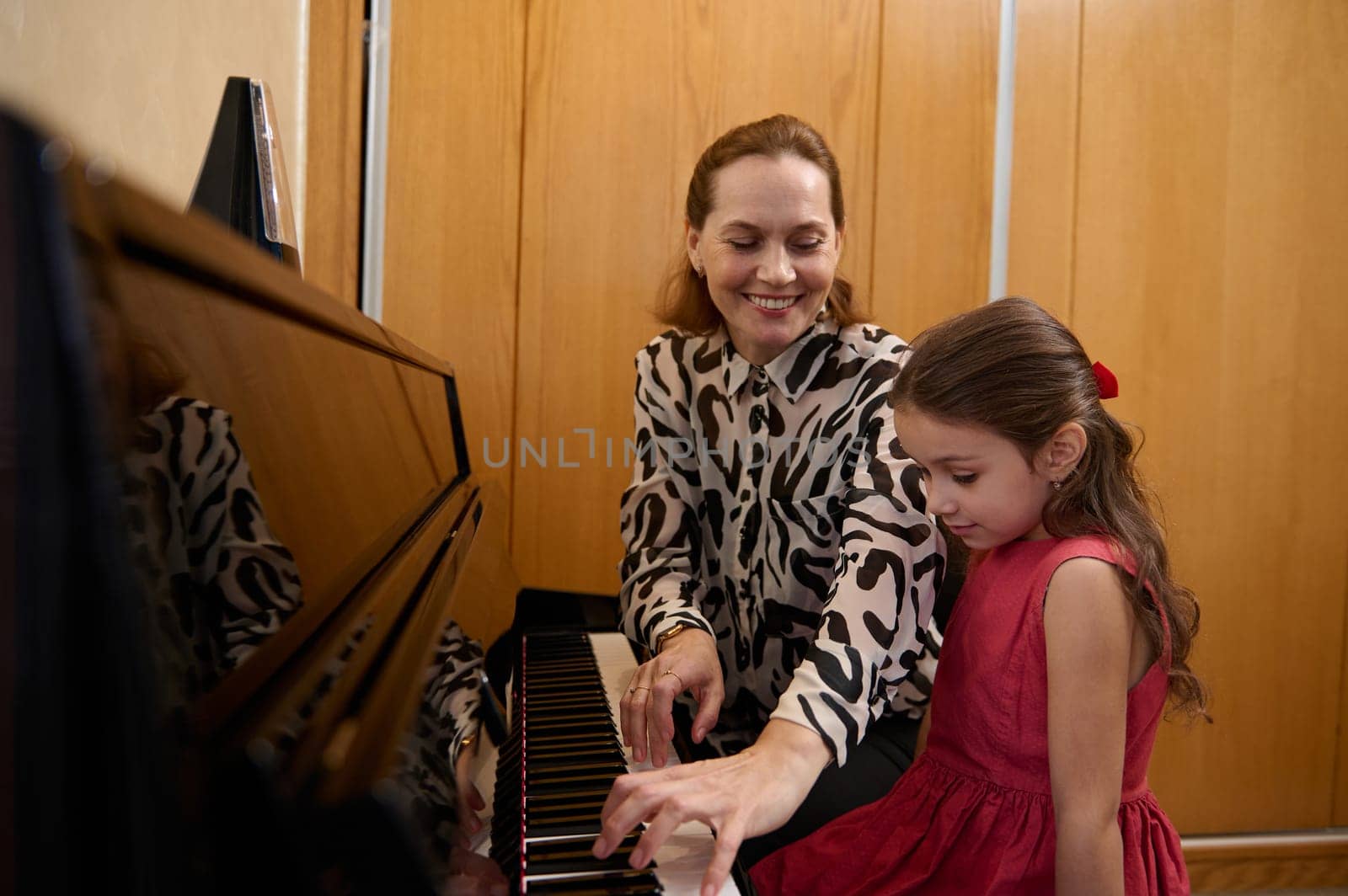 Cheerful beautiful woman pianist, musician teacher smiling while performing melody on piano, explaining music lesson to her student girl. Mother and daughter playing piano together
