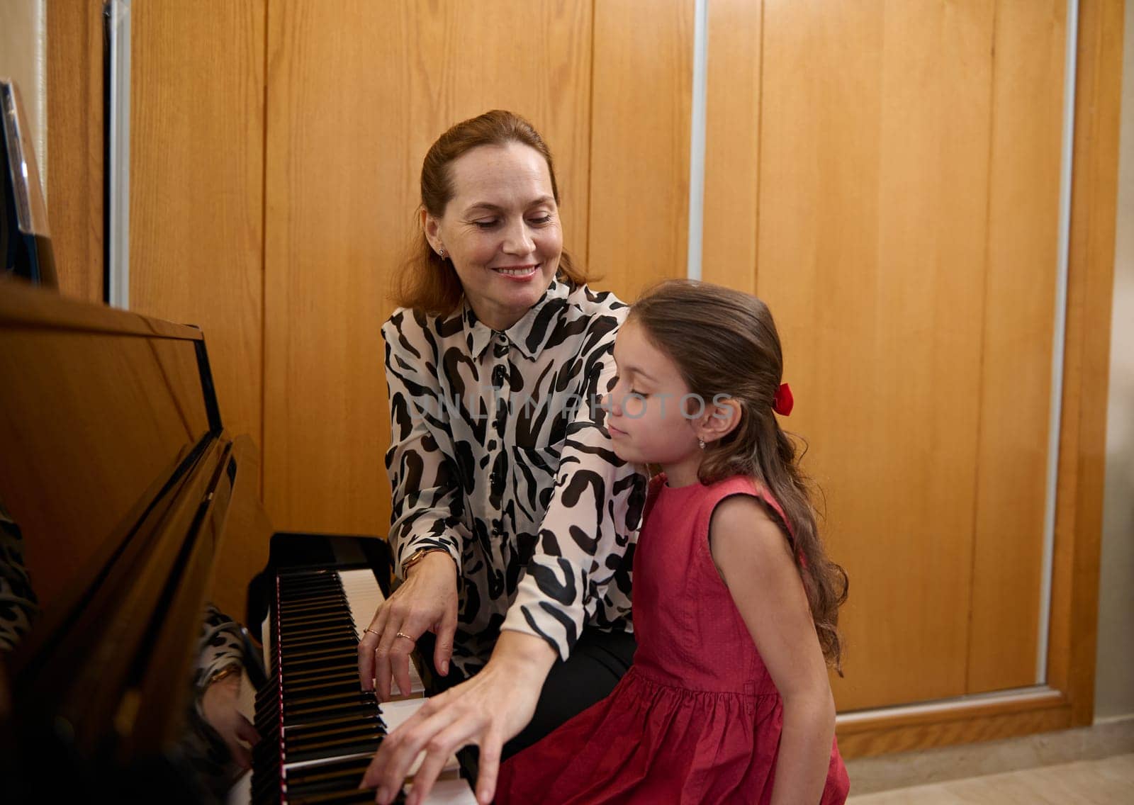 Cheerful mother and daughter pianists playing piano together, performing a melody for Christmas during music lesson together. Cute baby girl in elegant red dress, listens to her mom playing pianoforte by artgf