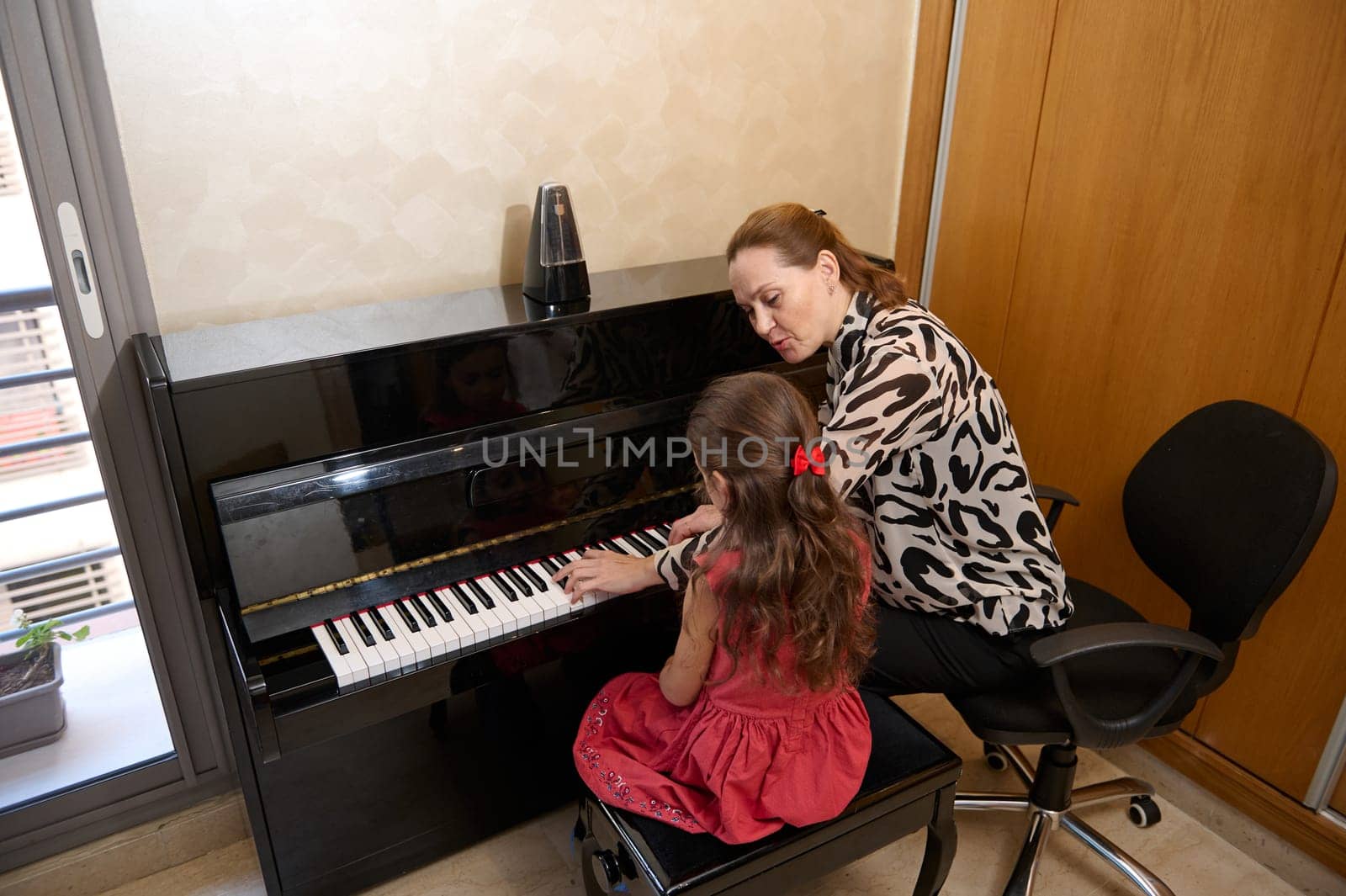 Musical education and talent development in progress. Rear view of a cute little girl in elegant red dress, sitting at wooden piano, with teacher learning to play piano indoors.