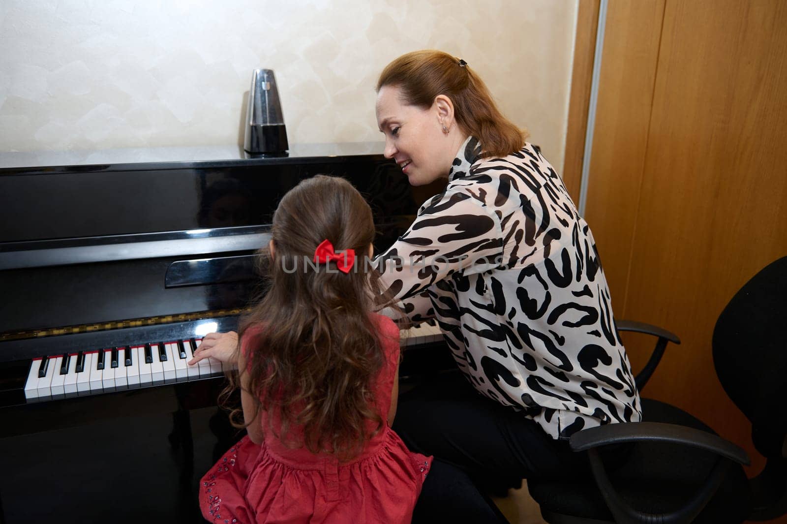 Inspired happy woman pianist, music teacher performing melody on piano forte, explaining piano lesson to a little girl, sitting nearby on a stool. Musical education and talent development in progress