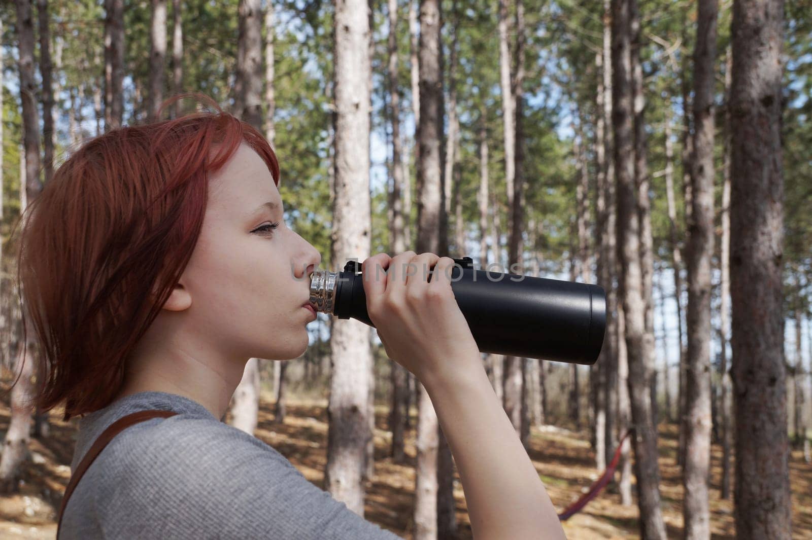 red-haired teenage girl drinks from a black thermos in the forest, side view