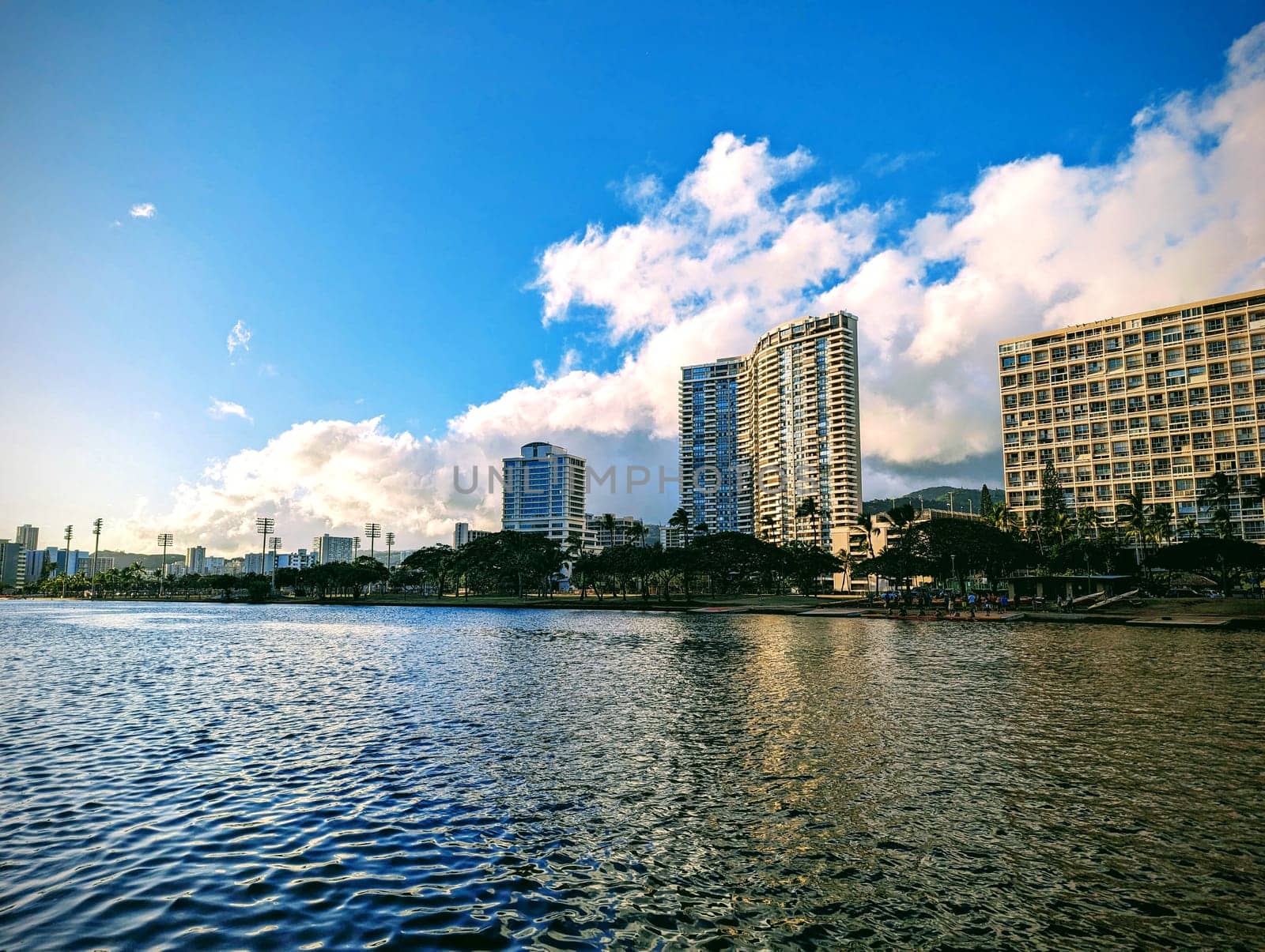 Serene banks of the Ala Wai Canal, an artificial waterway in Honolulu, Hawaii. The canal, stretching 1.5 miles (2.4 km), serves as the northern boundary of the vibrant Waikiki tourist district. Against a backdrop of towering buildings, the canal’s tranquil waters mirror the sky, creating a harmonious blend of urban and natural elements. The fluffy clouds overhead seem to float effortlessly, mirroring the peaceful ripples below. As the sun sets, the city lights begin to twinkle, casting a warm glow on the water’s surface. Whether you’re a local seeking a moment of respite or a visitor capturing memories, the Ala Wai Canal offers a quiet oasis amidst the bustling cityscape.
