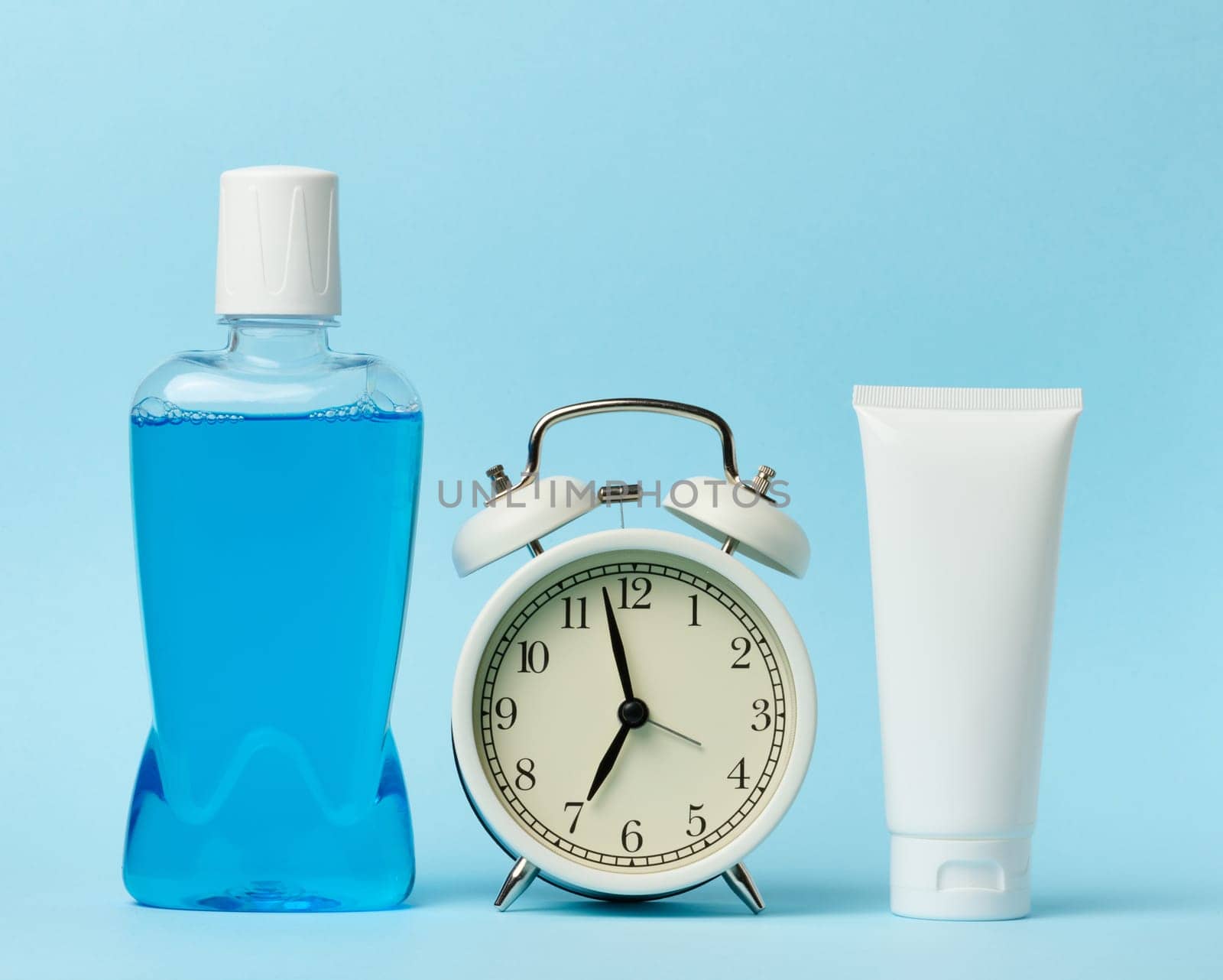 Plastic bottle with mouthwash, tube of toothpaste and round alarm clock, blue background